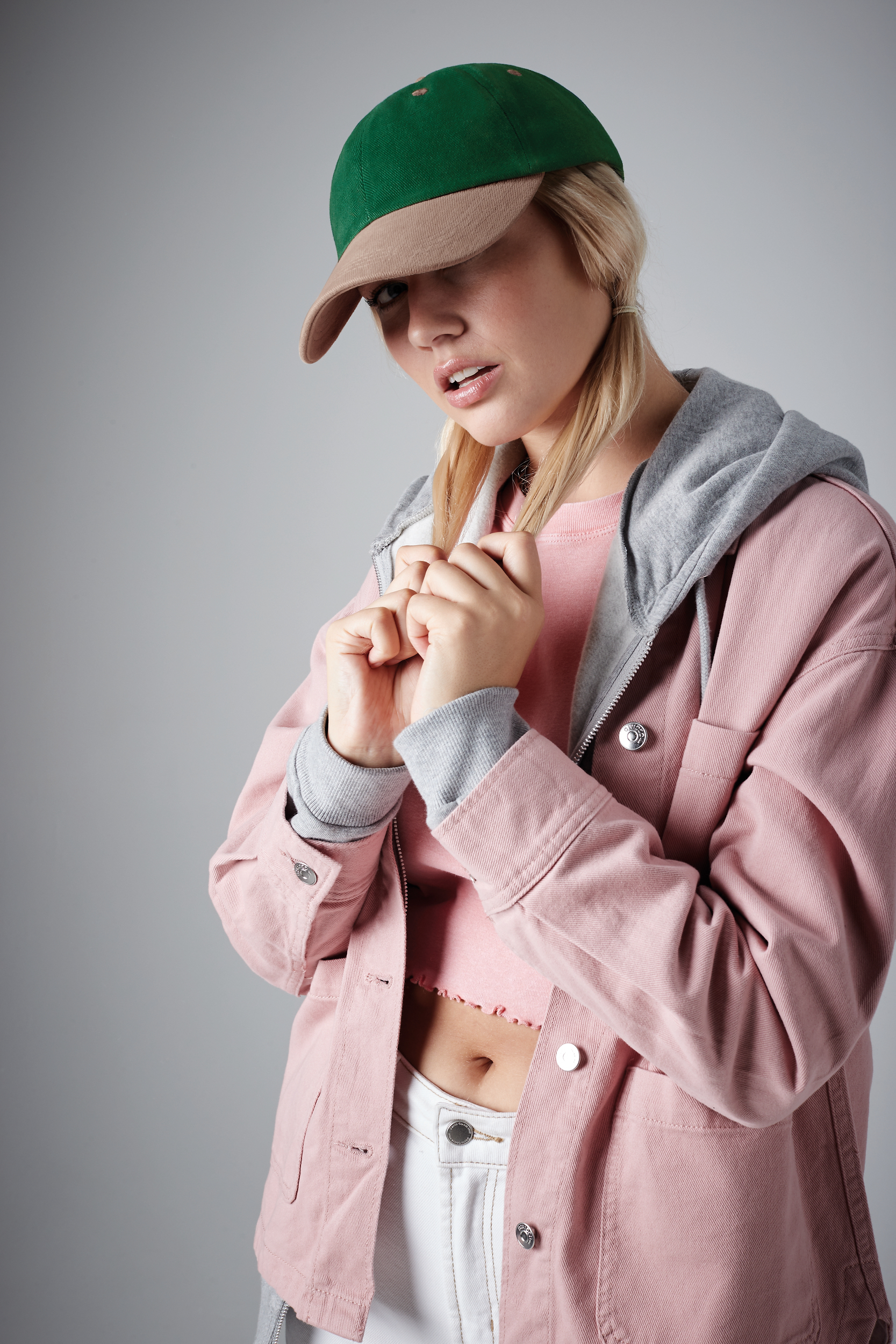 Mujer joven con gorra verde y beige, chaqueta rosa sobre sudadera gris y cinturilla blanca, sujetándose dos mechones de su pelo rubio sobre fondo neutro.