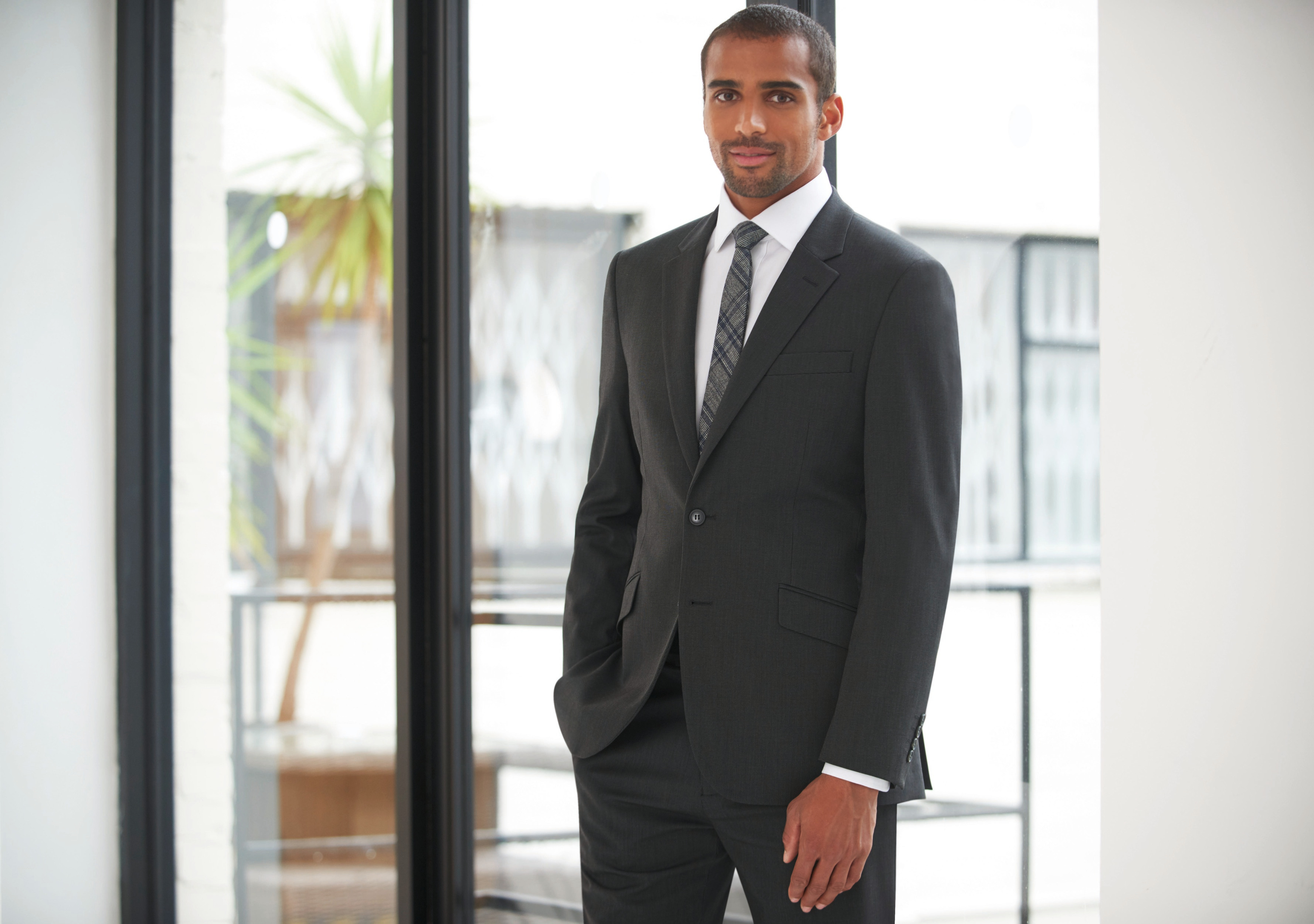 Hombre joven con traje formal de pie junto a un ventanal en una oficina moderna.