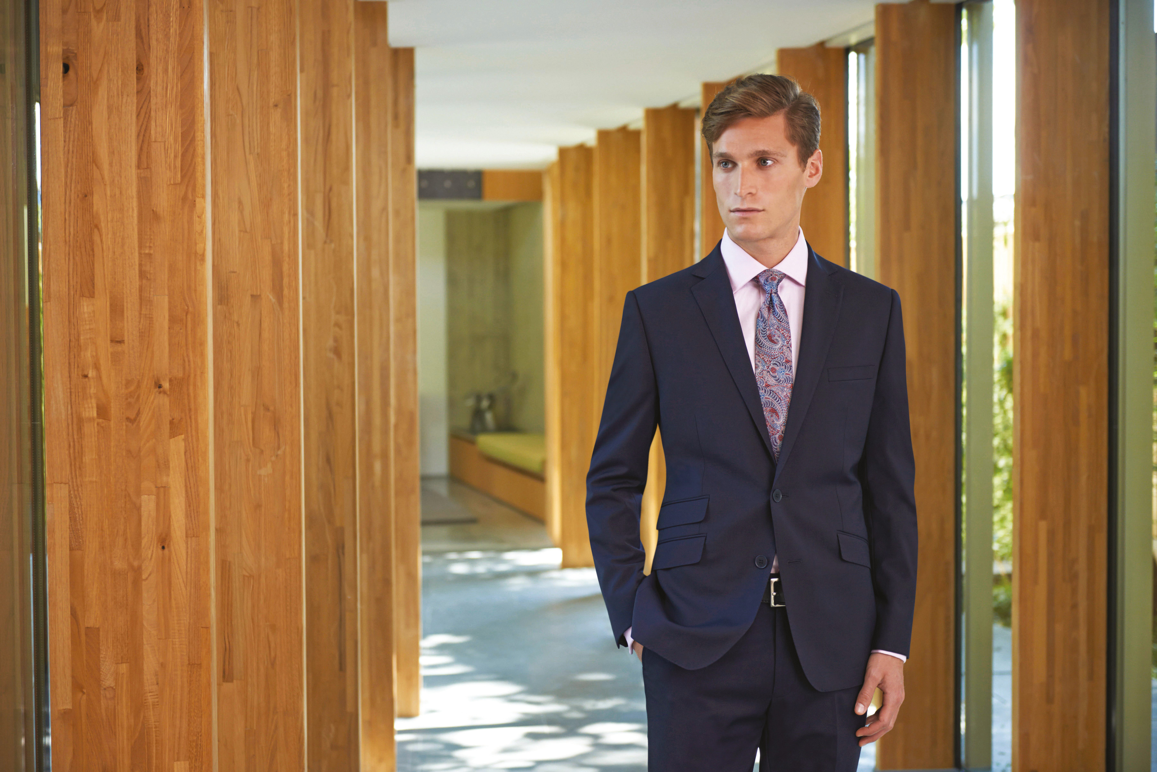 hombre joven con traje azul y corbata de estampado paisley posando en un pasillo con columnas de madera natural