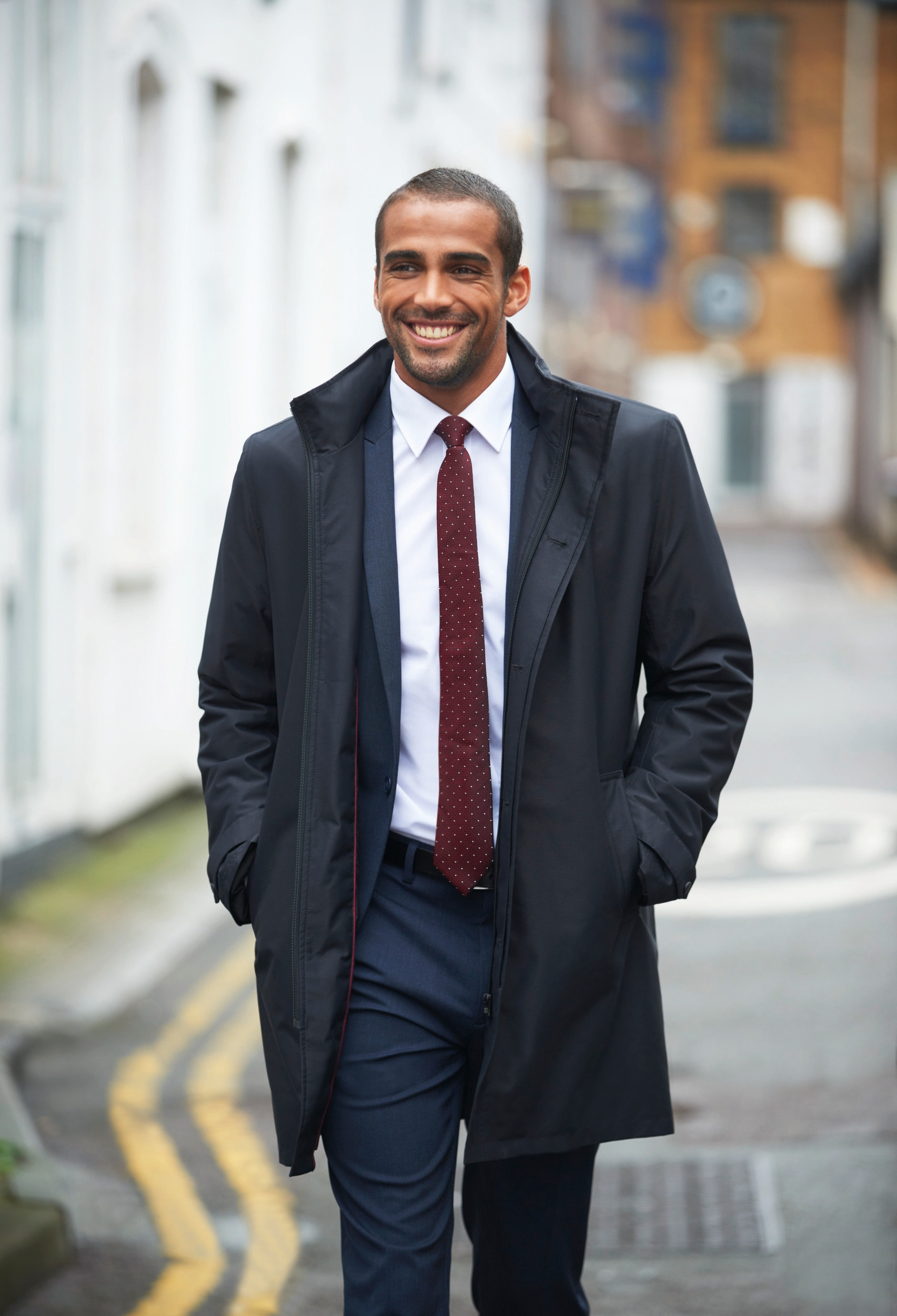 Hombre joven con traje, abrigo y corbata caminando sonriente por una calle urbana