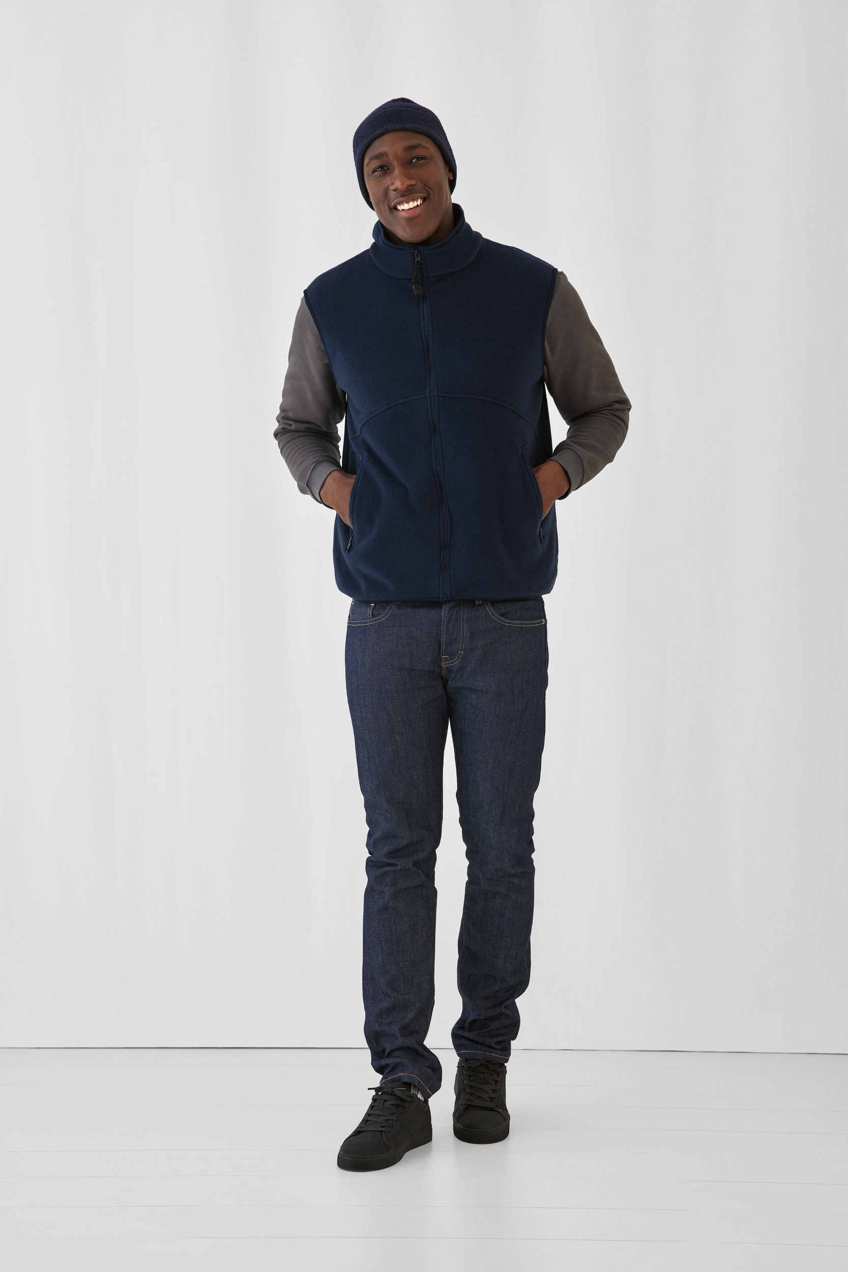 Hombre posando con chaleco polar azul marino, camiseta gris, vaqueros, gorro y zapatillas negras sobre fondo blanco