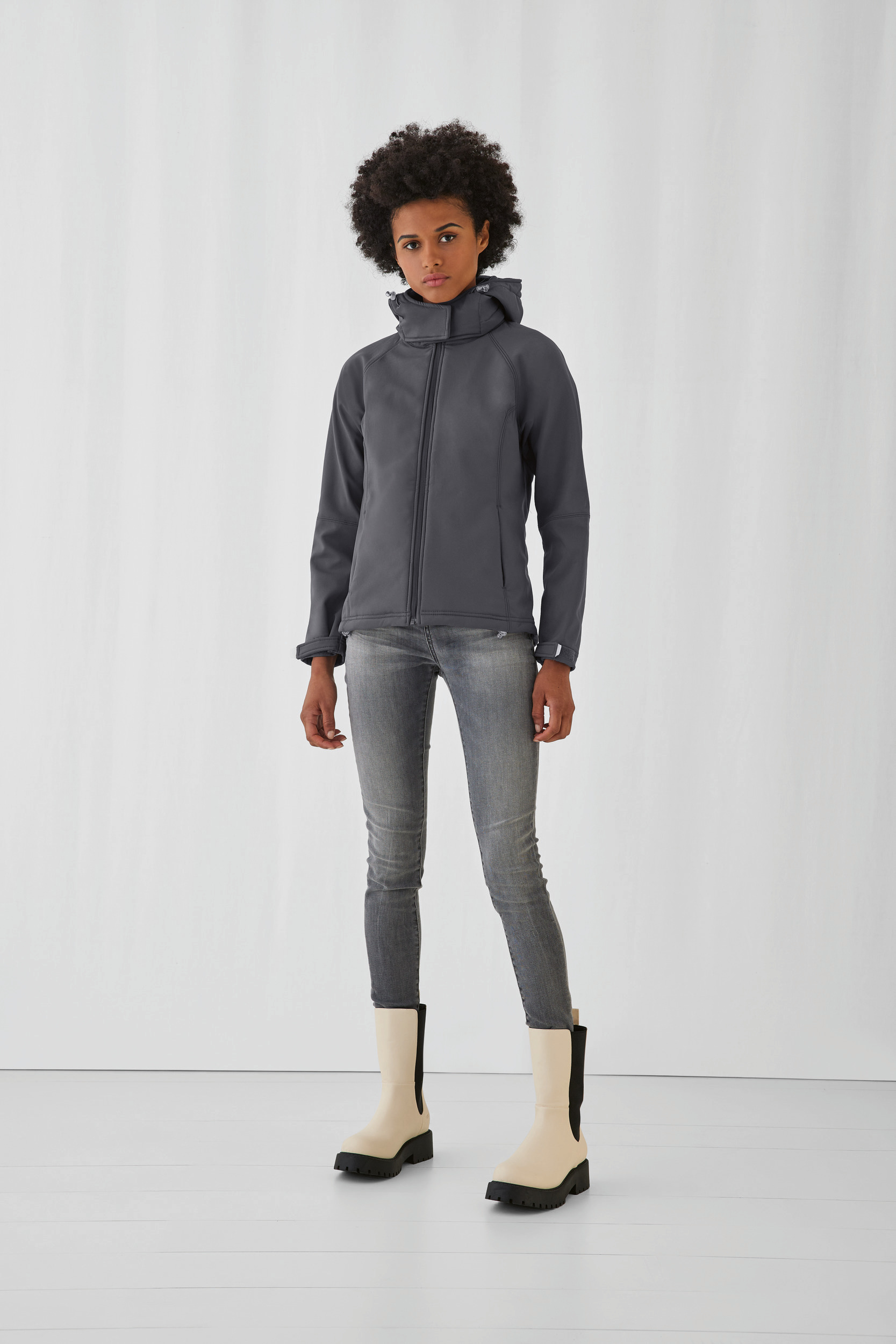 Mujer con chaqueta softshell gris, vaqueros ajustados y botas beige sobre fondo blanco