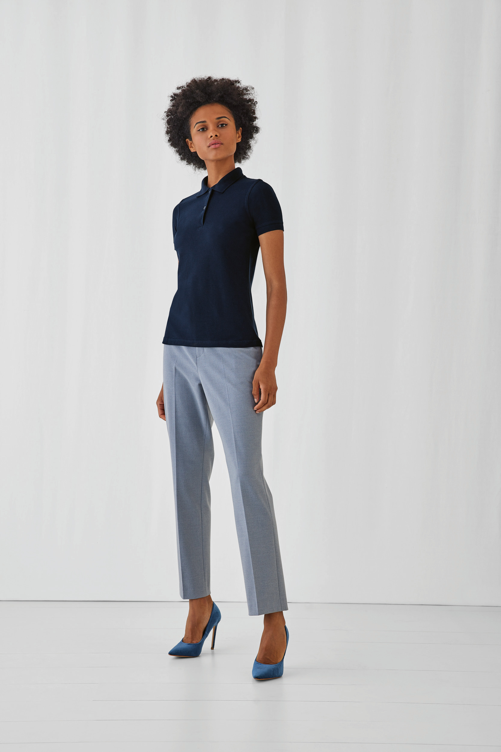 Mujer posando en estudio con polo azul marino, pantalones grises y zapatos de tacón azules.