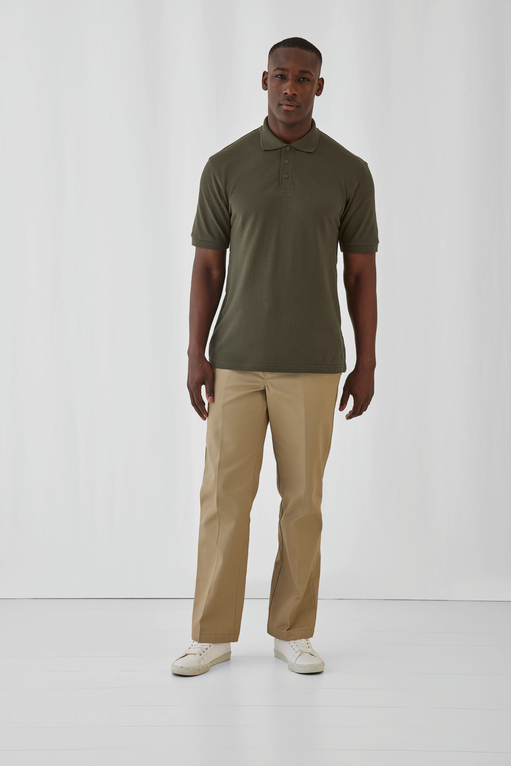 Hombre con polo verde oliva, pantalones beige y zapatillas blancas posando sobre fondo blanco.