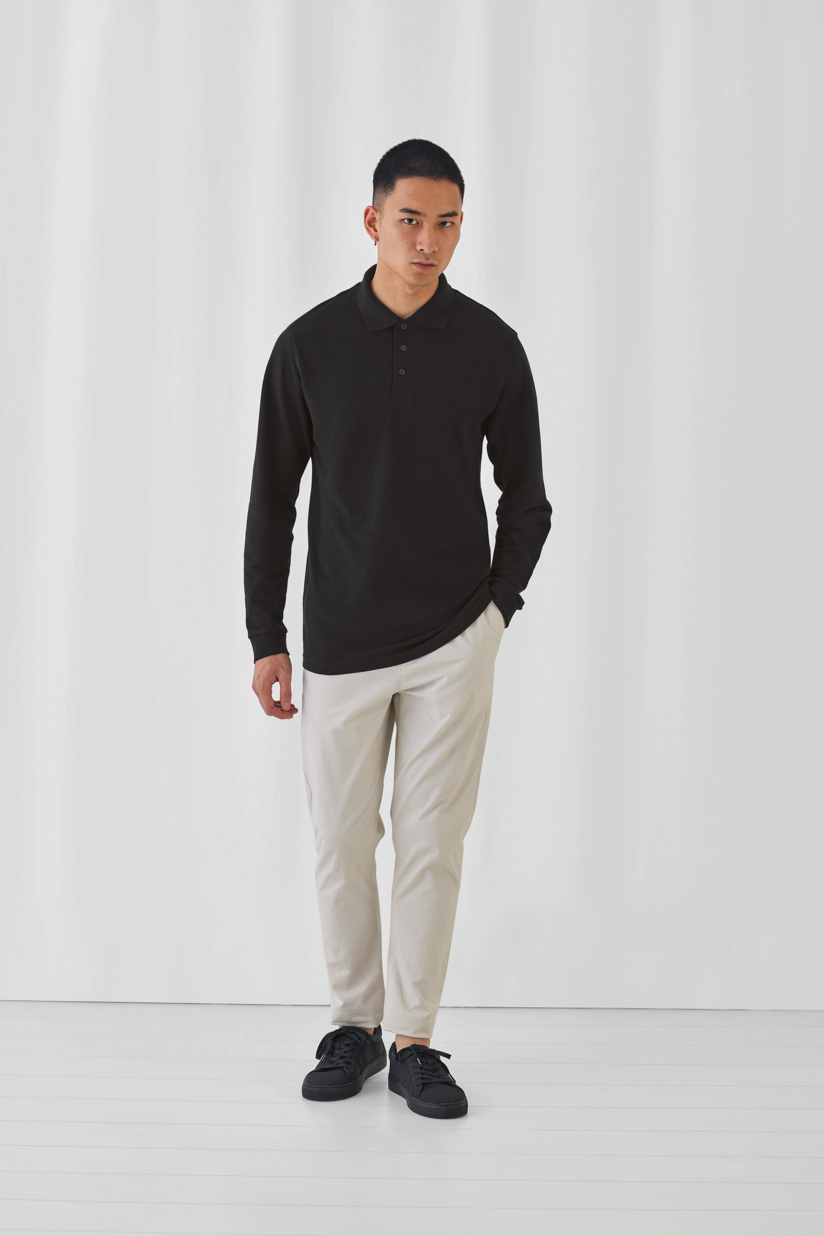 hombre joven de pie con polo de manga larga negro y pantalones chinos claros en fondo blanco