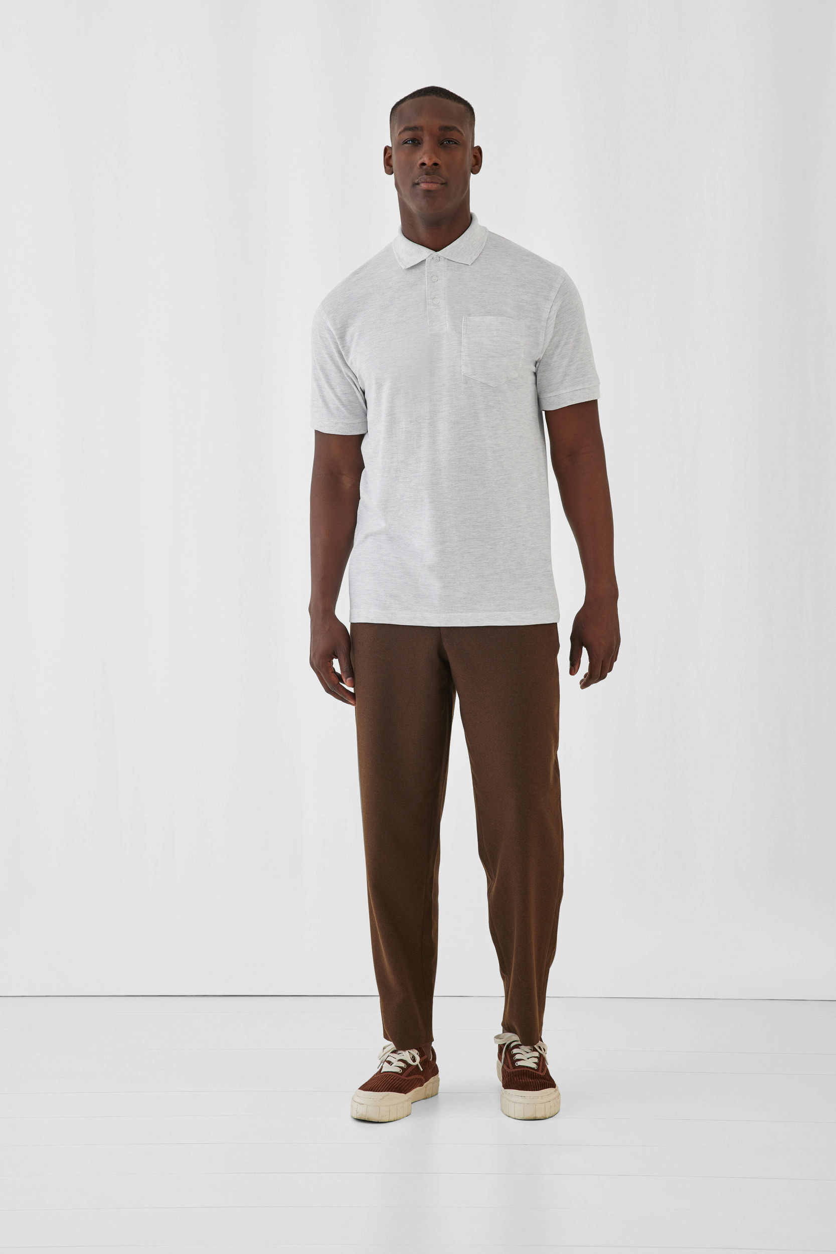 Hombre con polo gris de manga corta y pantalones marrones posando sobre fondo blanco.