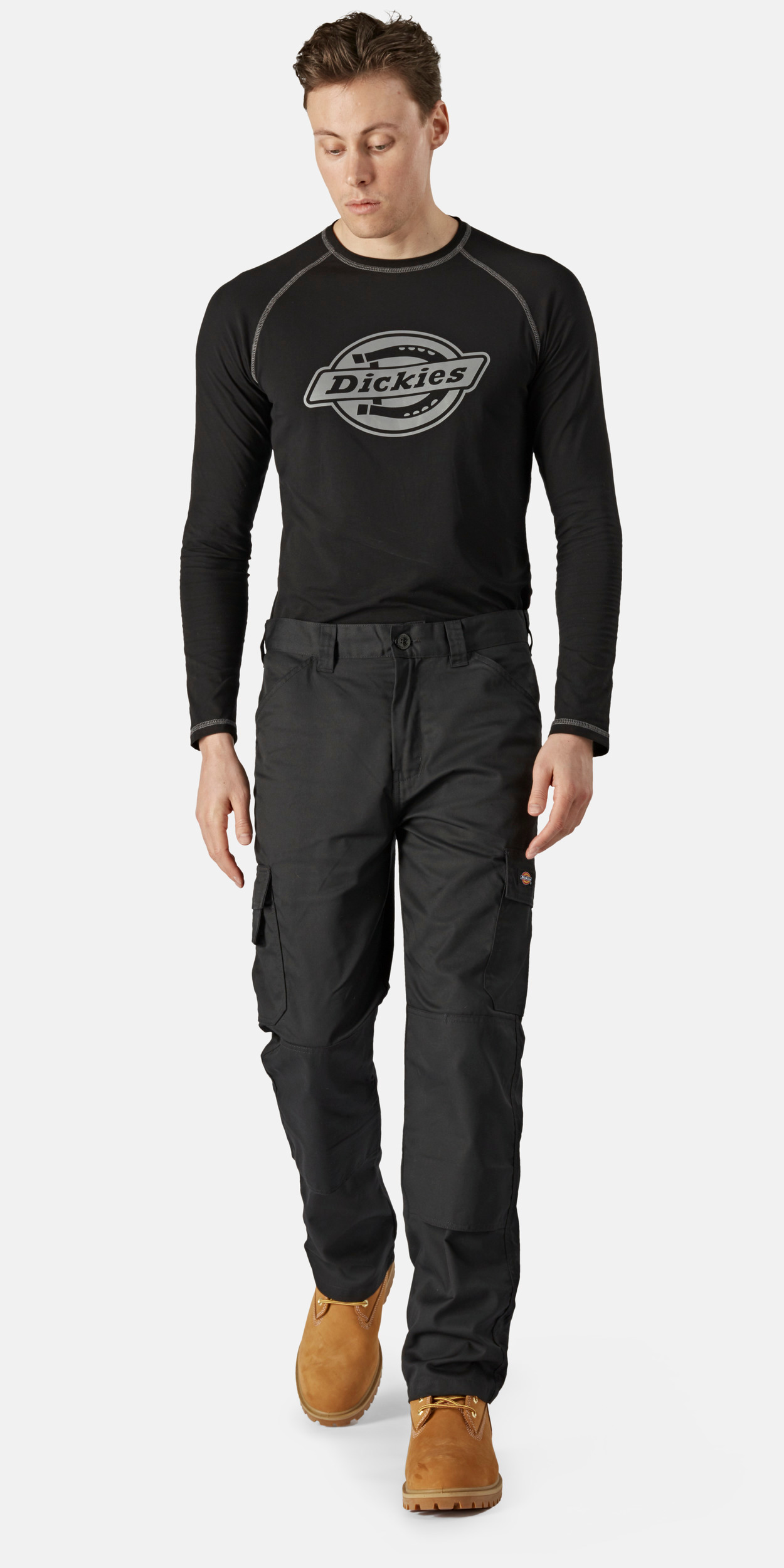 Hombre caminando con camiseta negra de manga larga con logo Dickies, pantalones cargo negros y botas de trabajo marrones sobre fondo blanco.