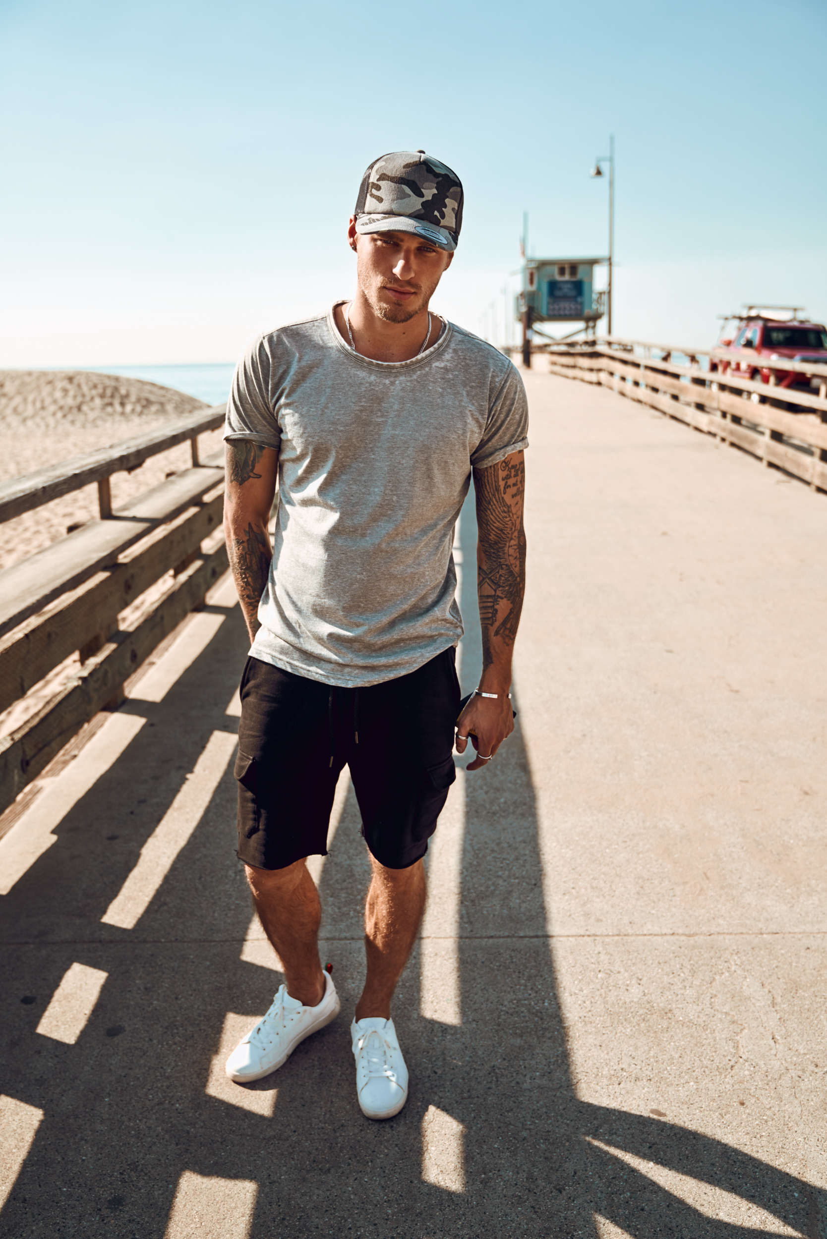 hombre con gorra de camuflaje y tatuajes de pie en un muelle junto a la playa bajo el sol