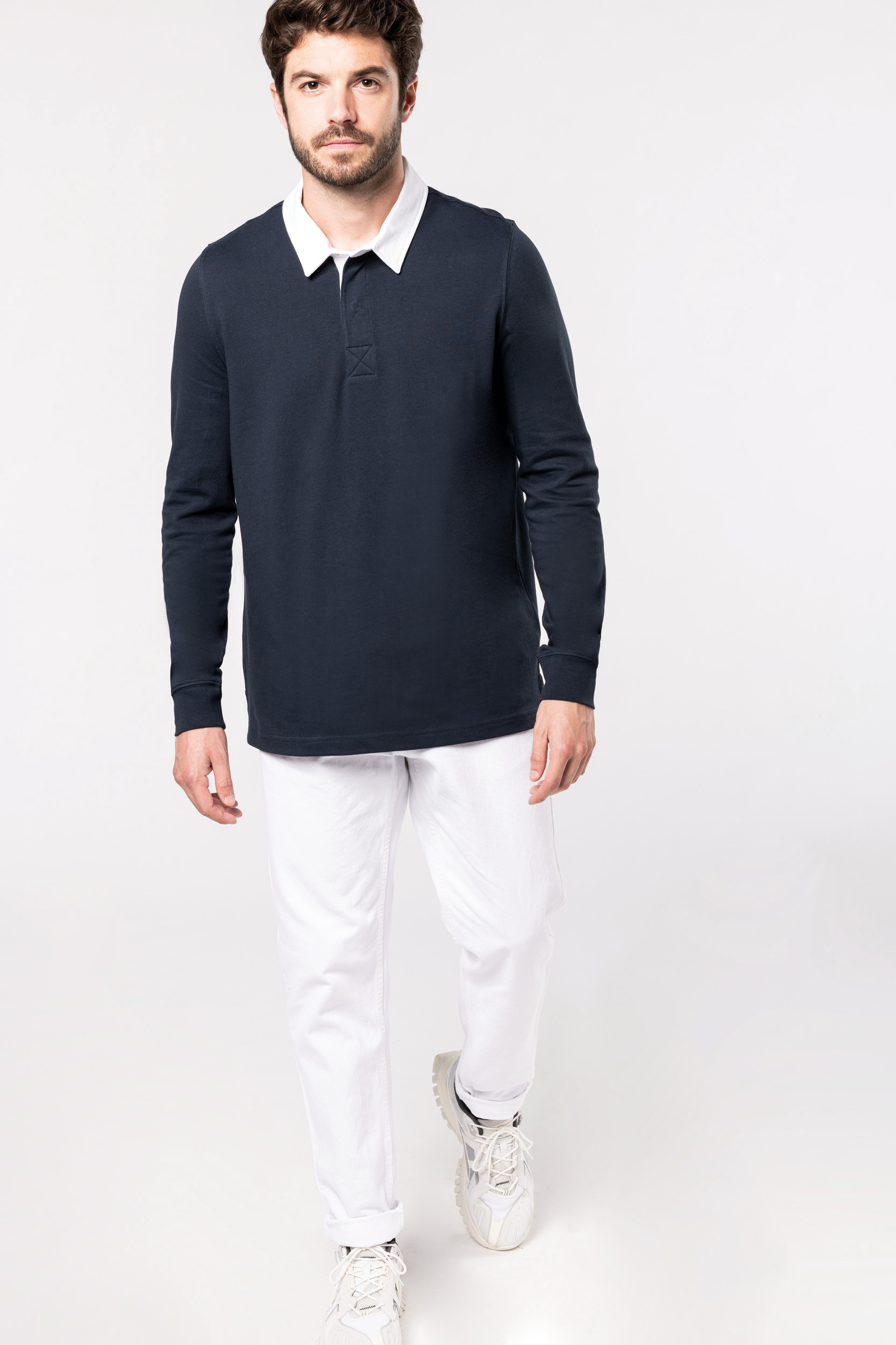 Hombre caminando sobre fondo blanco con polo azul marino de manga larga, pantalones blancos y zapatillas deportivas claras