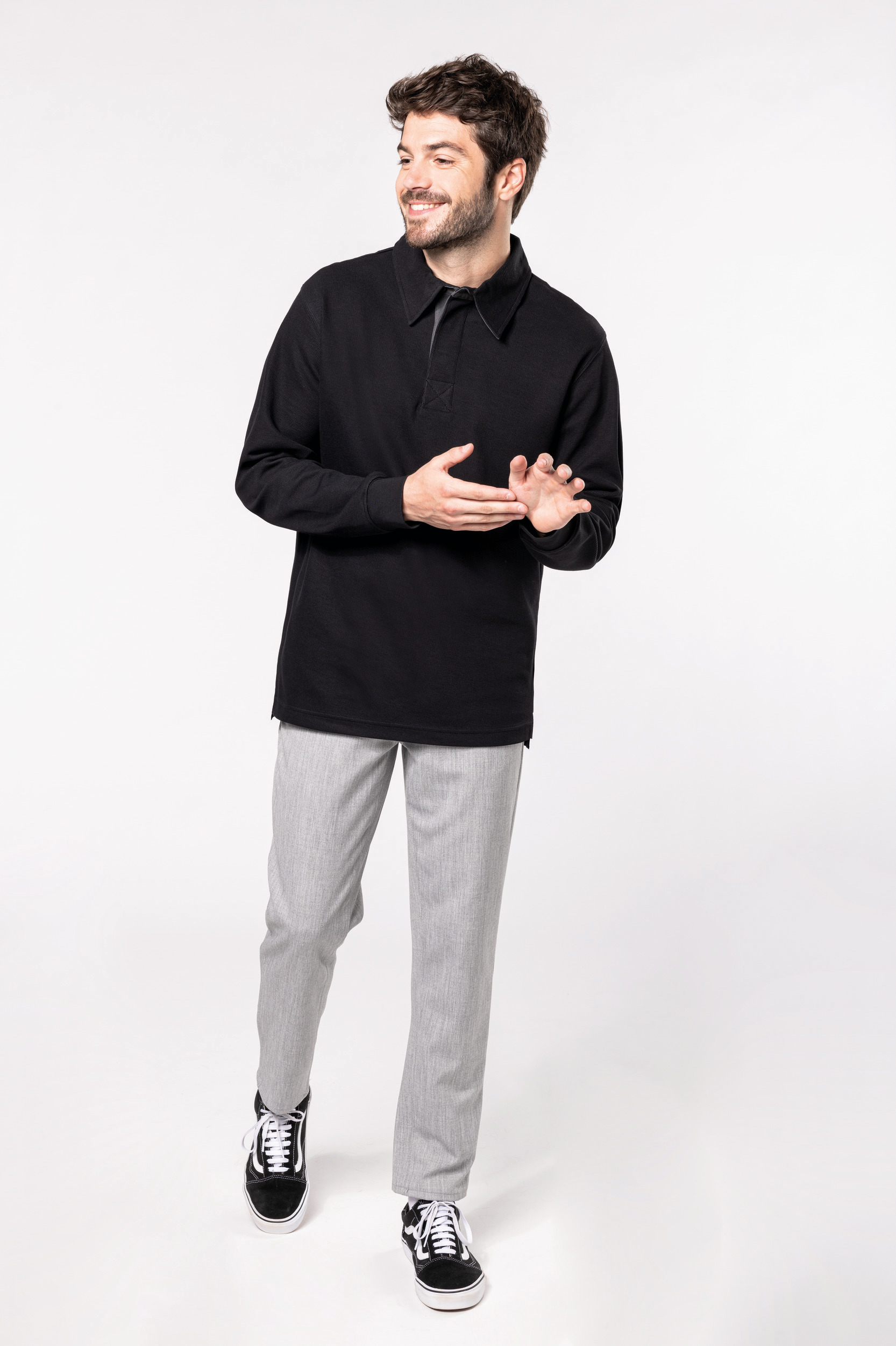 Hombre joven sonriente con polo negro de manga larga, pantalones grises y zapatillas negras sobre fondo blanco.