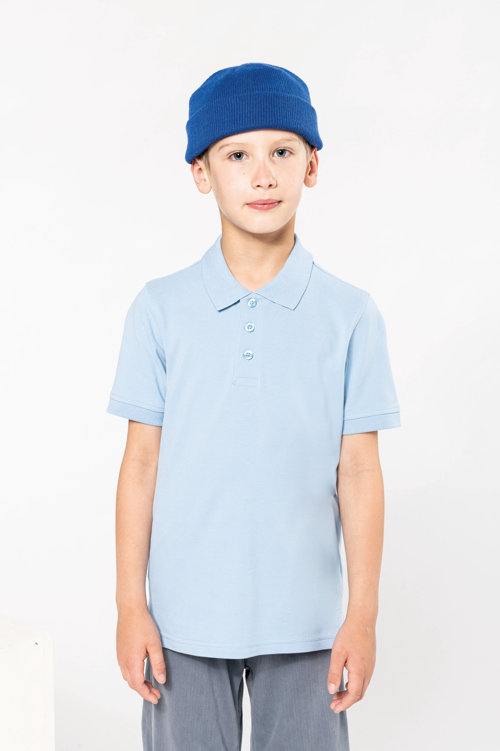 Niño de pie con gorro de punto azul y polo celeste sobre fondo blanco.