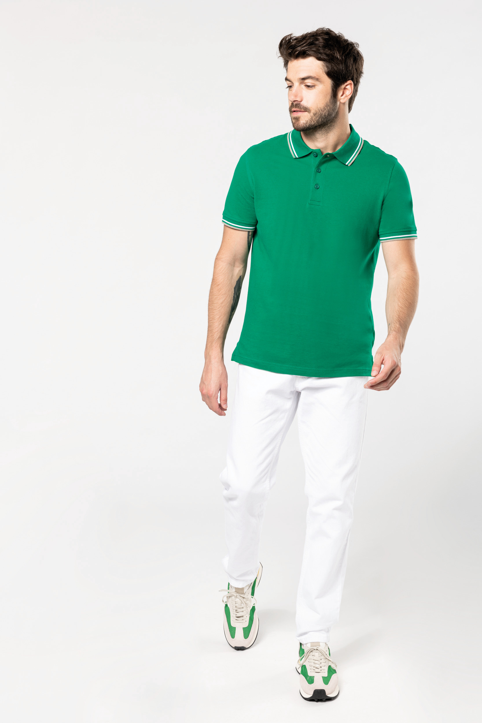 Hombre joven caminando con polo verde, vaqueros blancos y zapatillas deportivas a juego sobre fondo claro