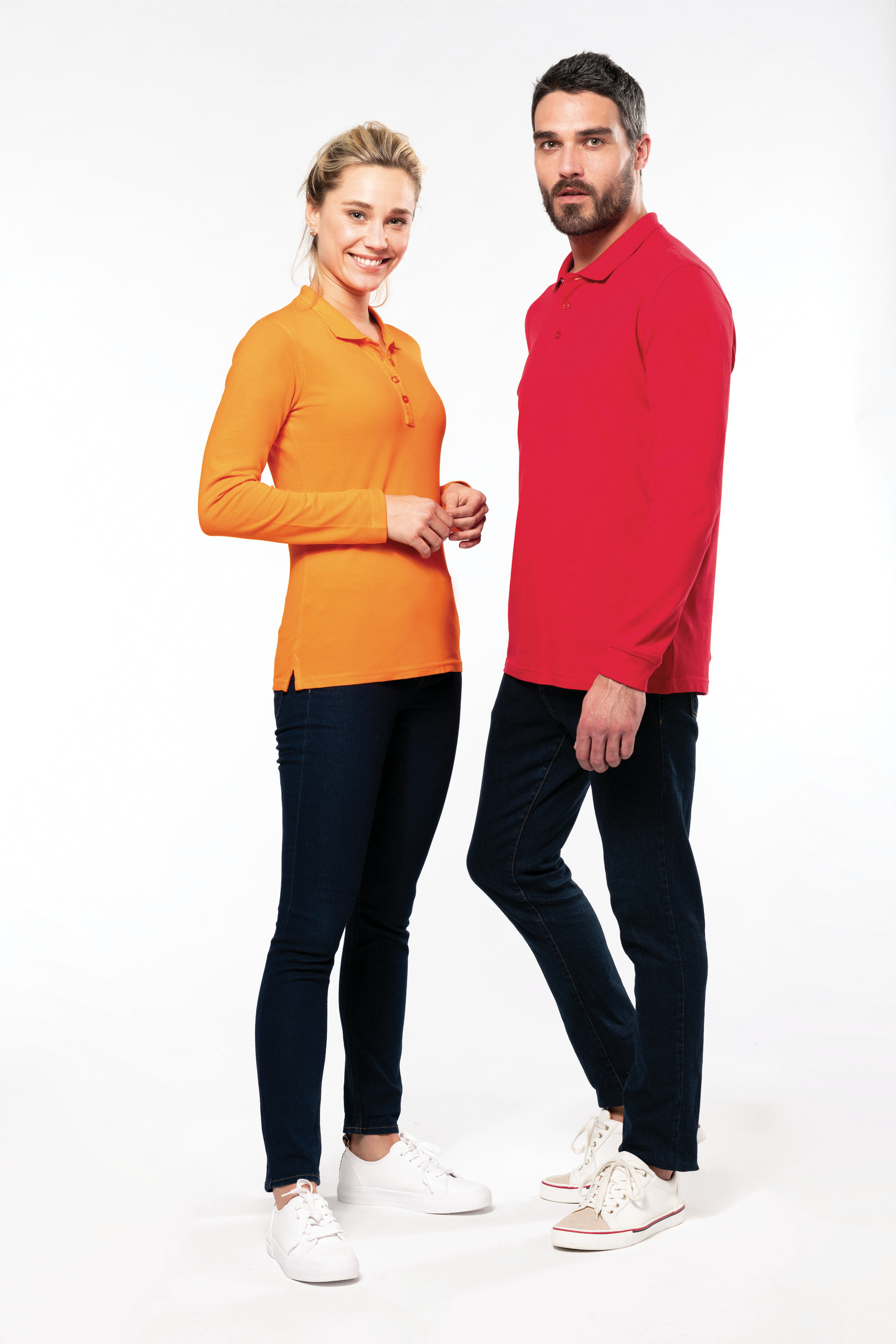 Pareja de modelos de pie en estudio con fondo blanco, ella viste polo naranja y él polo rojo con vaqueros y zapatillas blancas.
