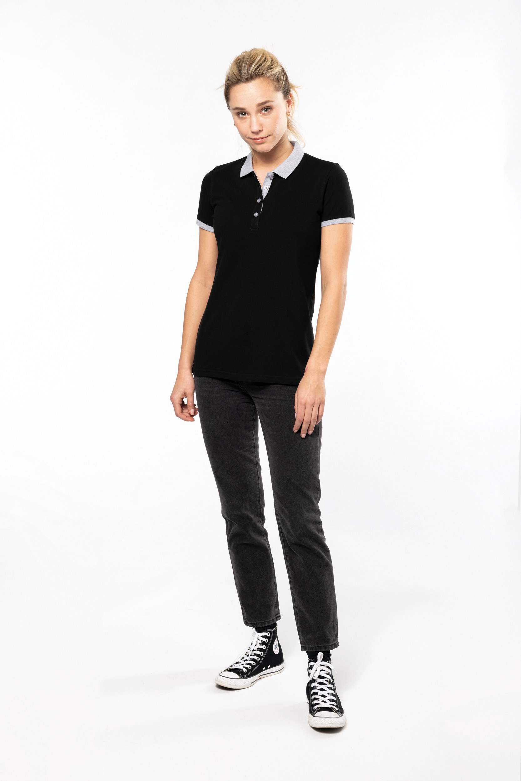 Mujer joven de pie sobre fondo blanco con polo negro de cuello gris, vaqueros oscuros y zapatillas negras
