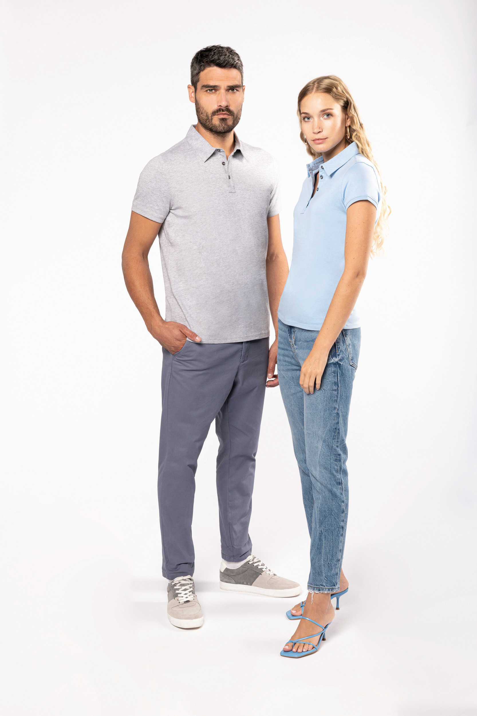 Hombre y mujer de pie sobre fondo blanco con polo gris y camiseta polo celeste en estilo casual.