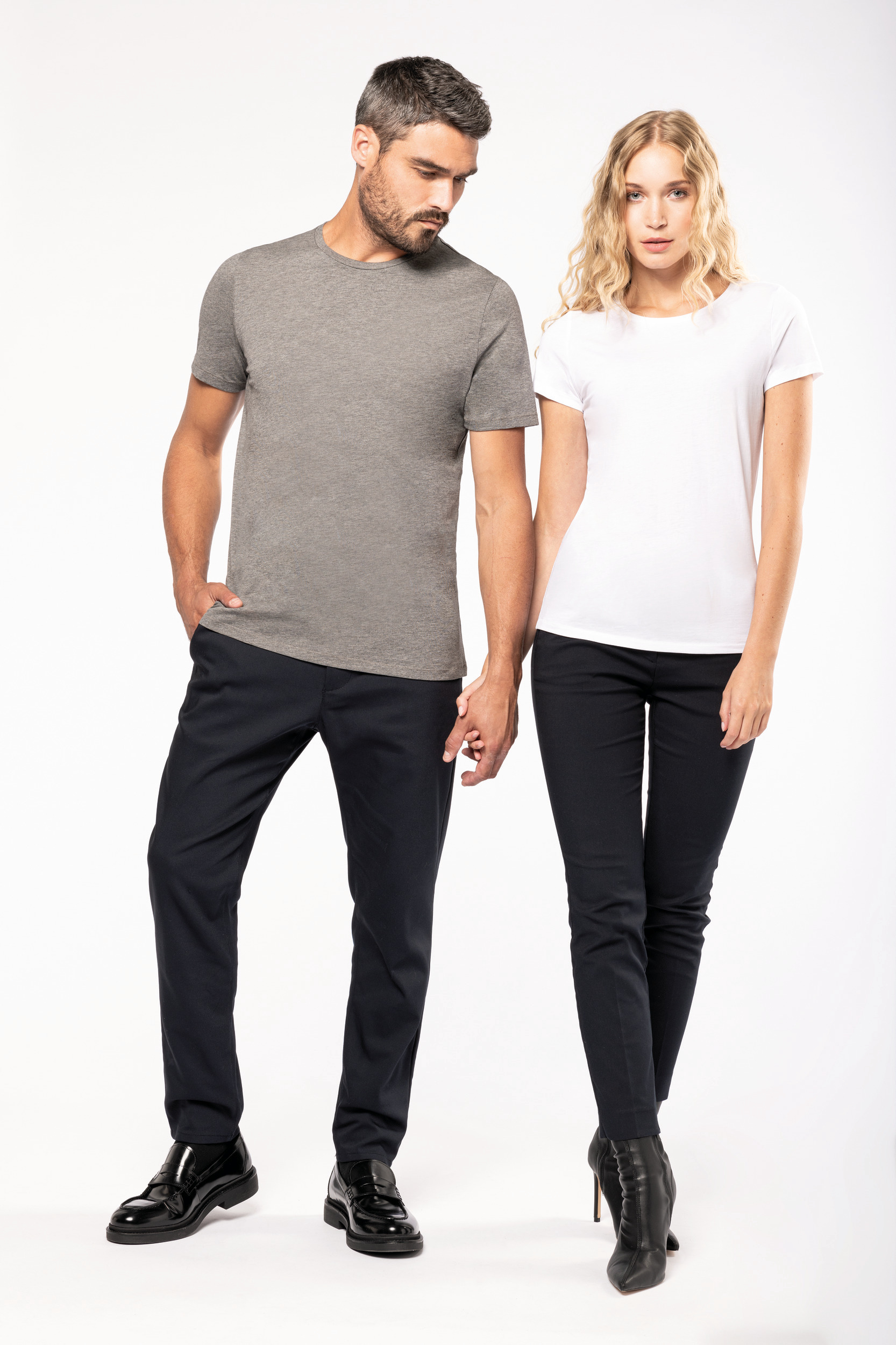 Pareja de modelos de pie con camisetas básicas y pantalones oscuros sonrientes y cogidos de la mano sobre fondo blanco