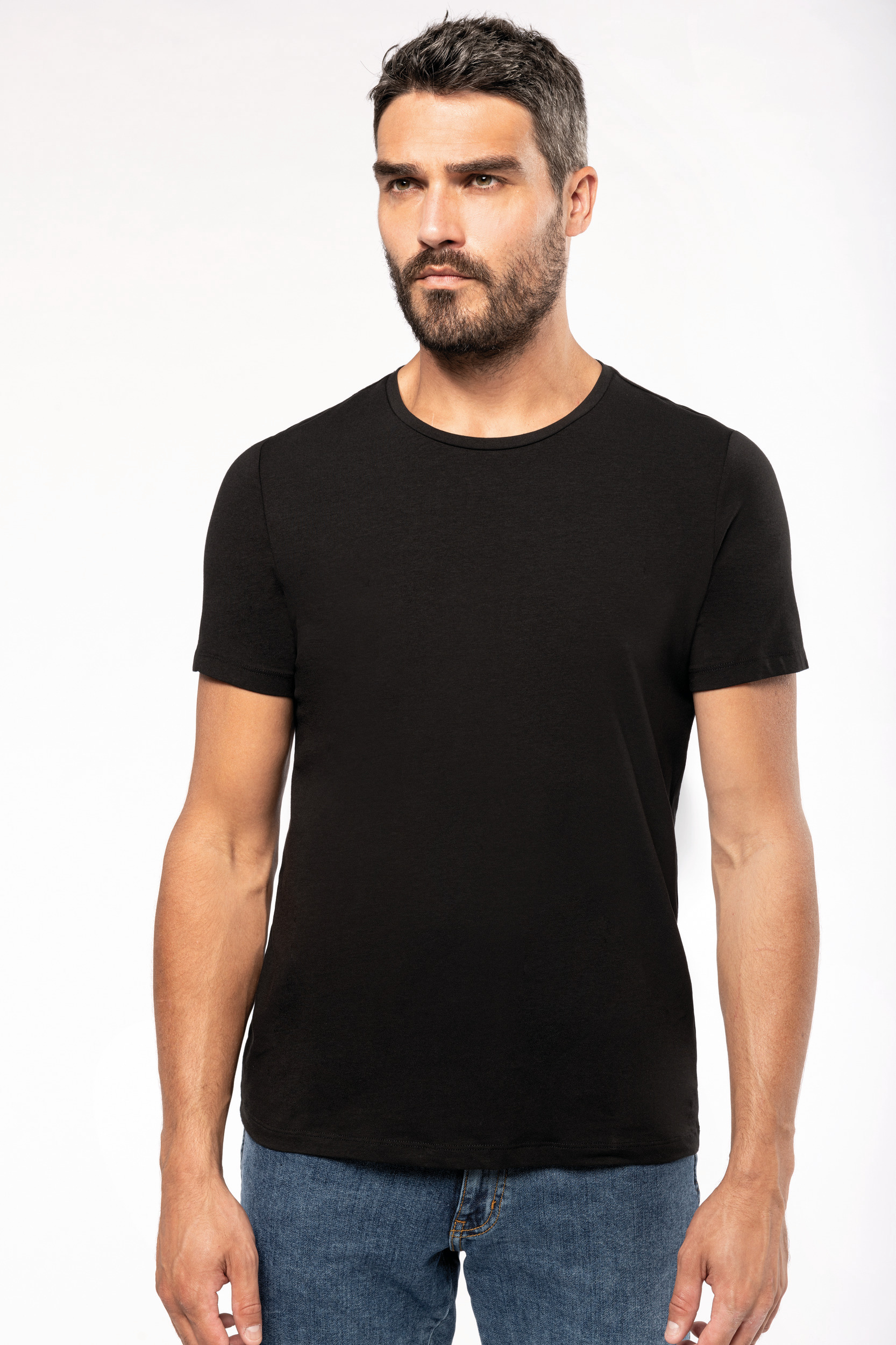 Hombre de pie con camiseta negra de manga corta y vaqueros, posando sobre fondo blanco.