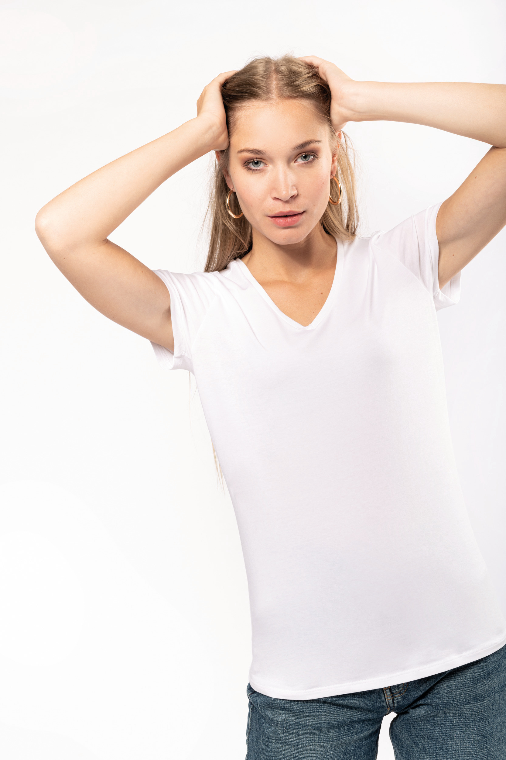 Mujer rubia con camiseta blanca y vaqueros posando sobre fondo blanco con las manos en la cabeza
