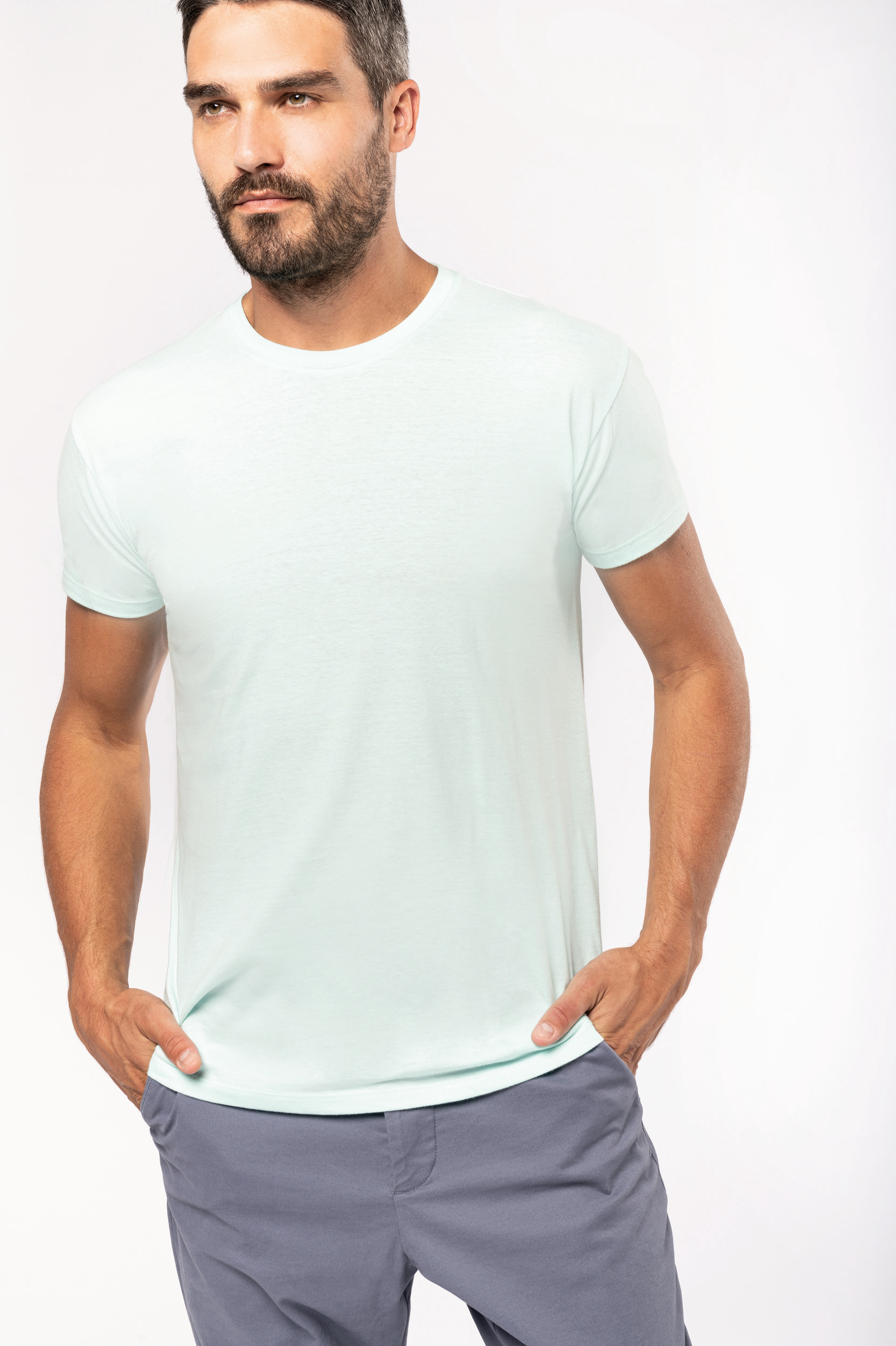 hombre con camiseta blanca y pantalones grises de pie con las manos en los bolsillos sobre fondo neutro