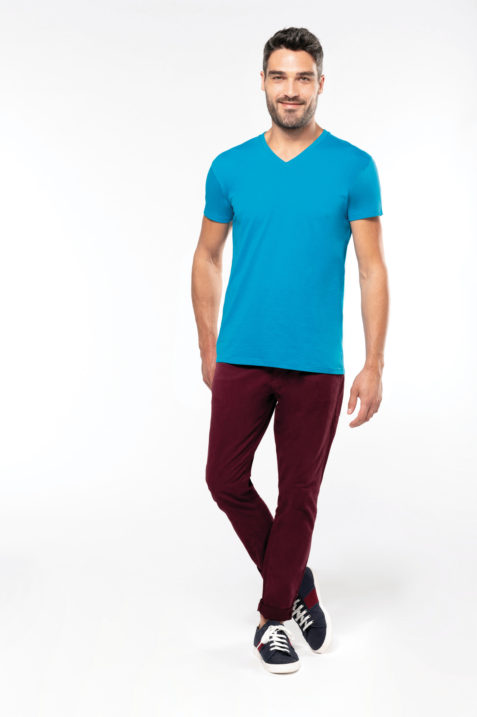 Hombre joven de pie con camiseta azul, pantalones granate y zapatillas sobre fondo blanco
