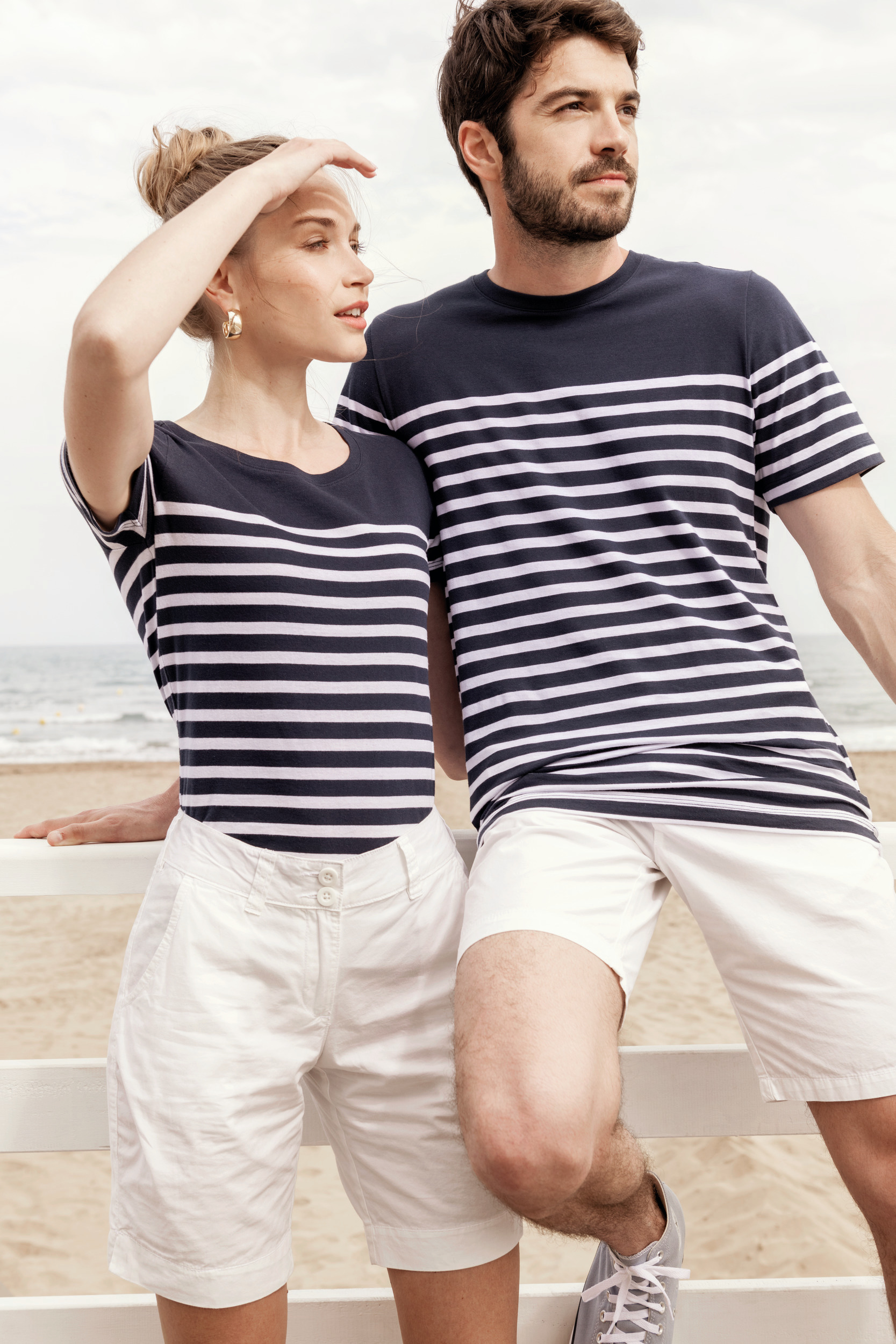 Pareja apoyada en una barandilla frente al mar con camiseta de rayas marineras y pantalones blancos.