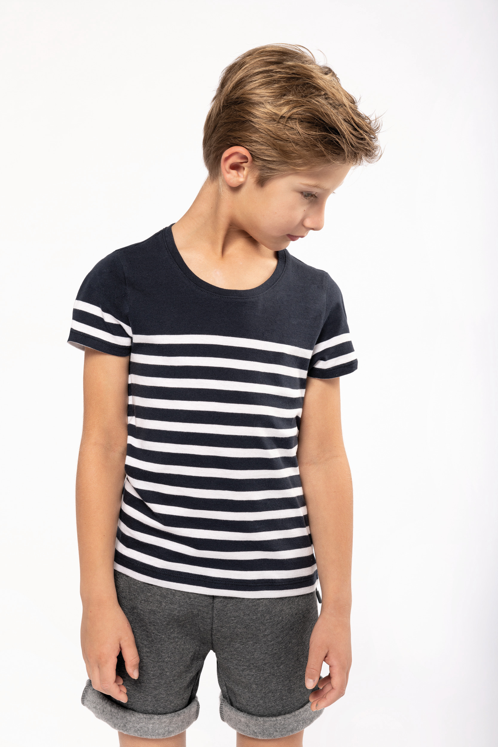 Niño con camiseta de rayas azul y blanca y pantalones cortos grises mirando hacia abajo sobre fondo blanco.