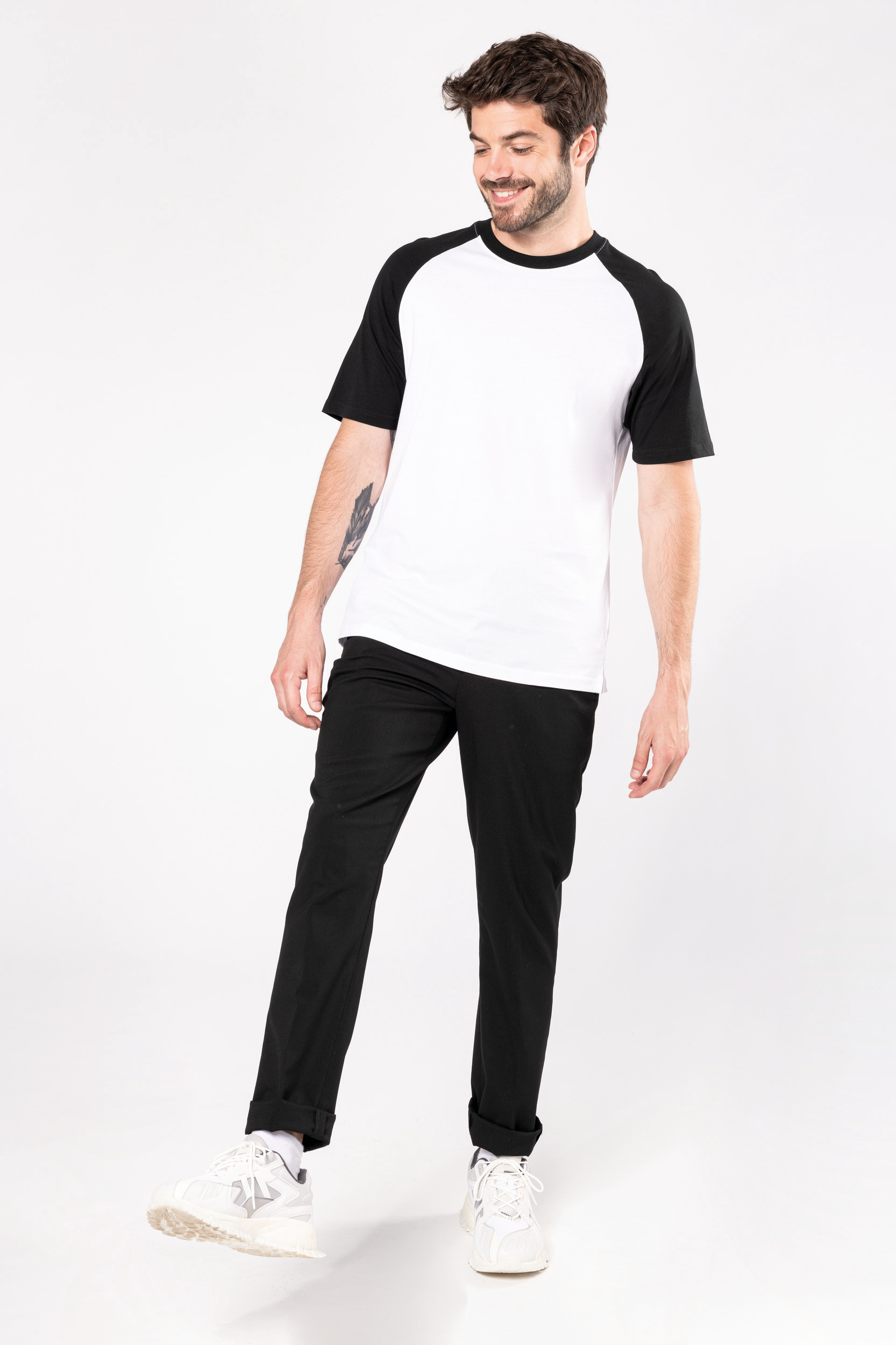 Hombre joven sonriente con camiseta blanca de mangas negras y pantalones negros sobre fondo blanco.