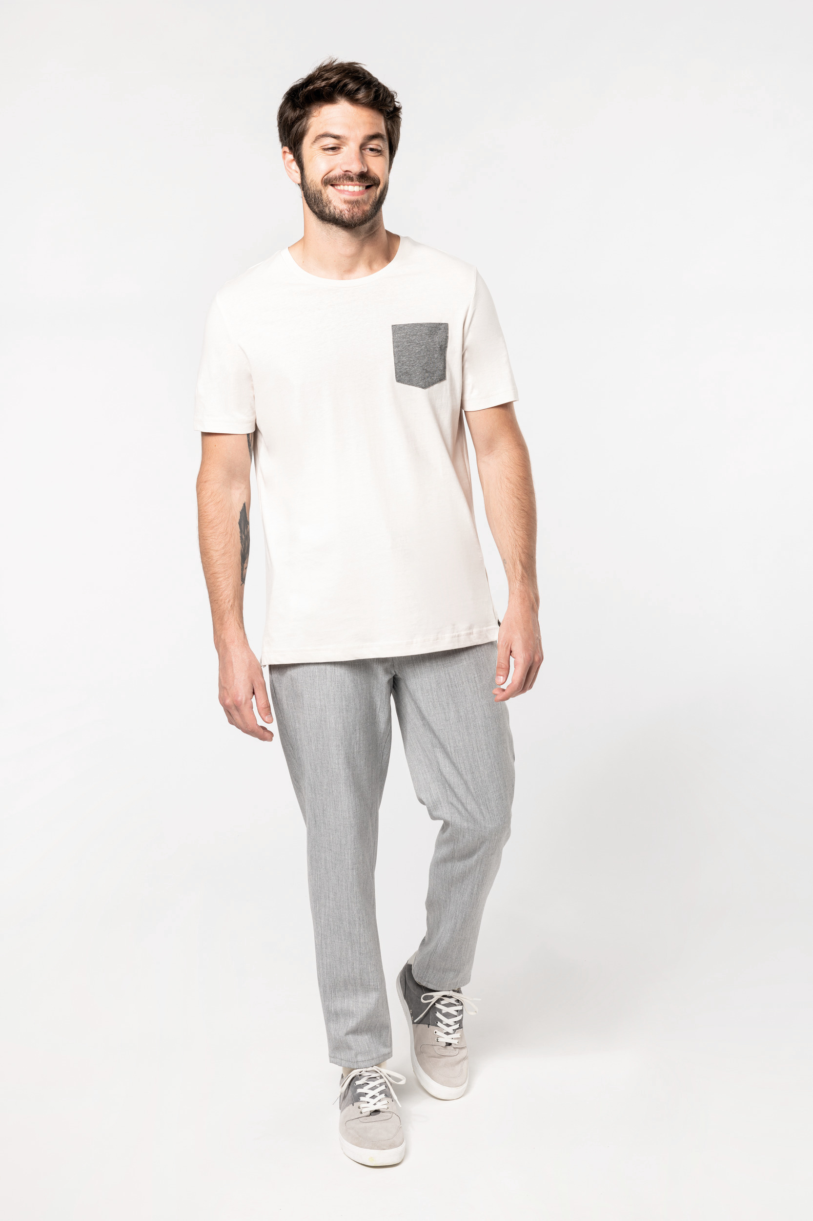 Hombre joven sonriente caminando con camiseta blanca de bolsillo gris, pantalones y zapatillas grises sobre fondo blanco.