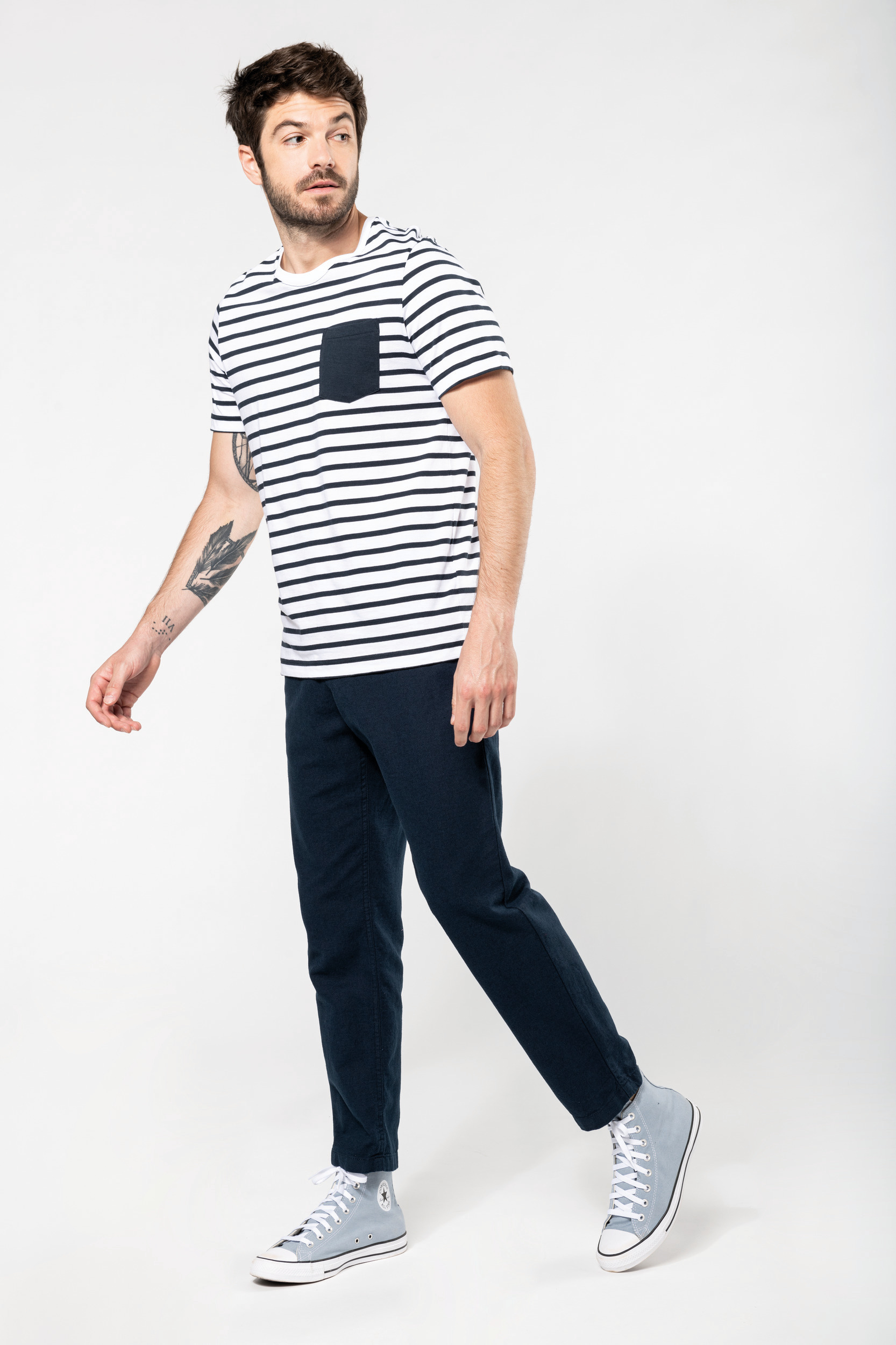 Hombre joven con camiseta de rayas marineras, pantalones azul marino y zapatillas altas grises sobre fondo blanco.
