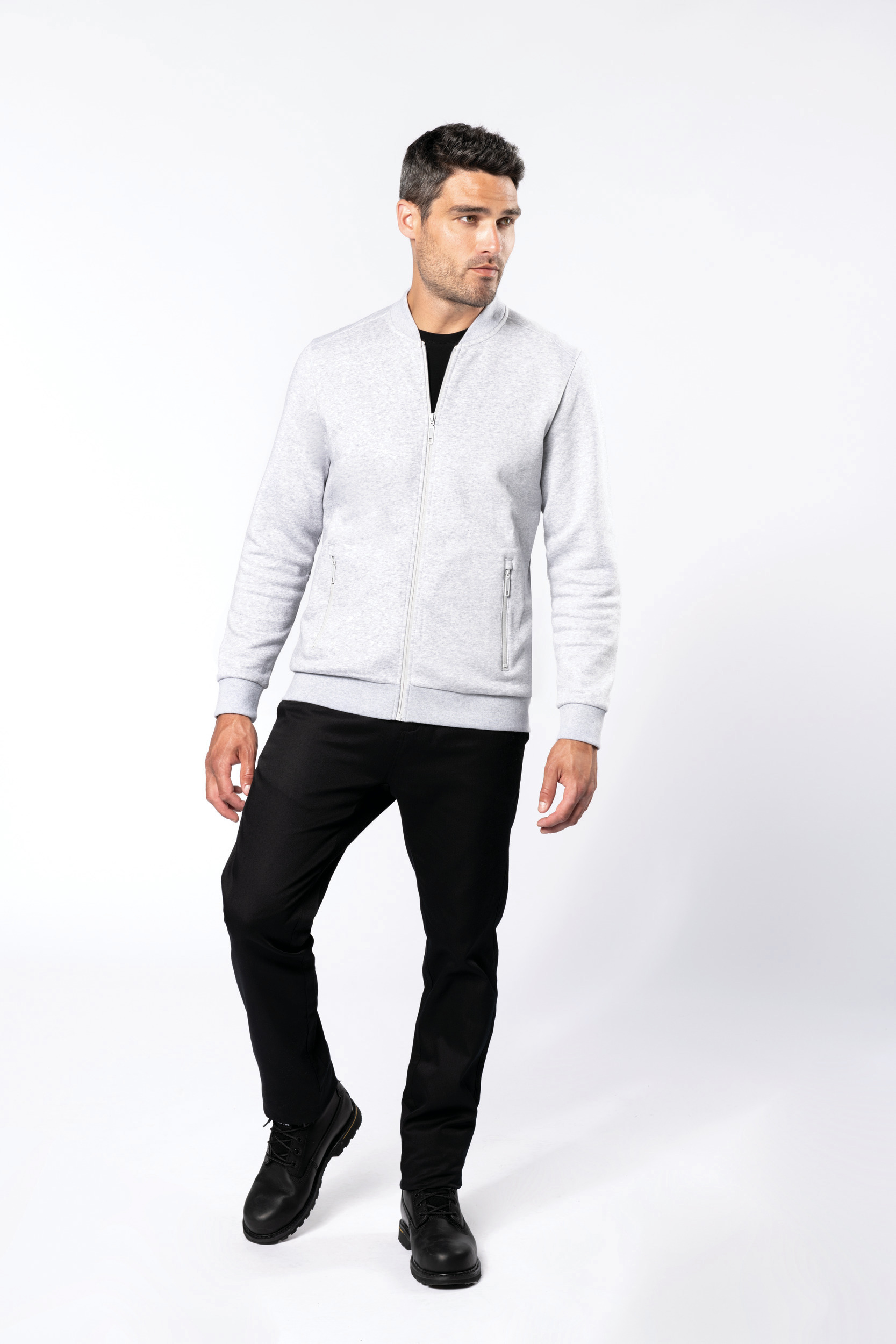Hombre joven de pie con chaqueta gris de cremallera, pantalones negros y botas sobre fondo blanco