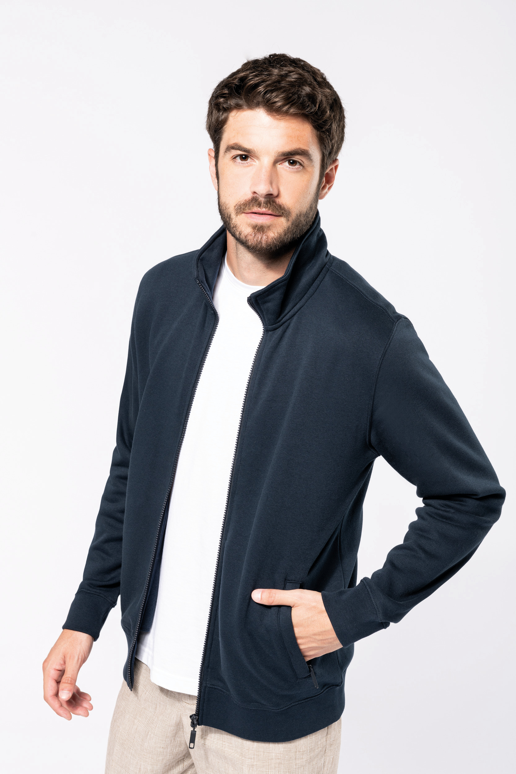 Hombre joven de pie con chaqueta azul marino de cremallera y camiseta blanca sobre fondo blanco.