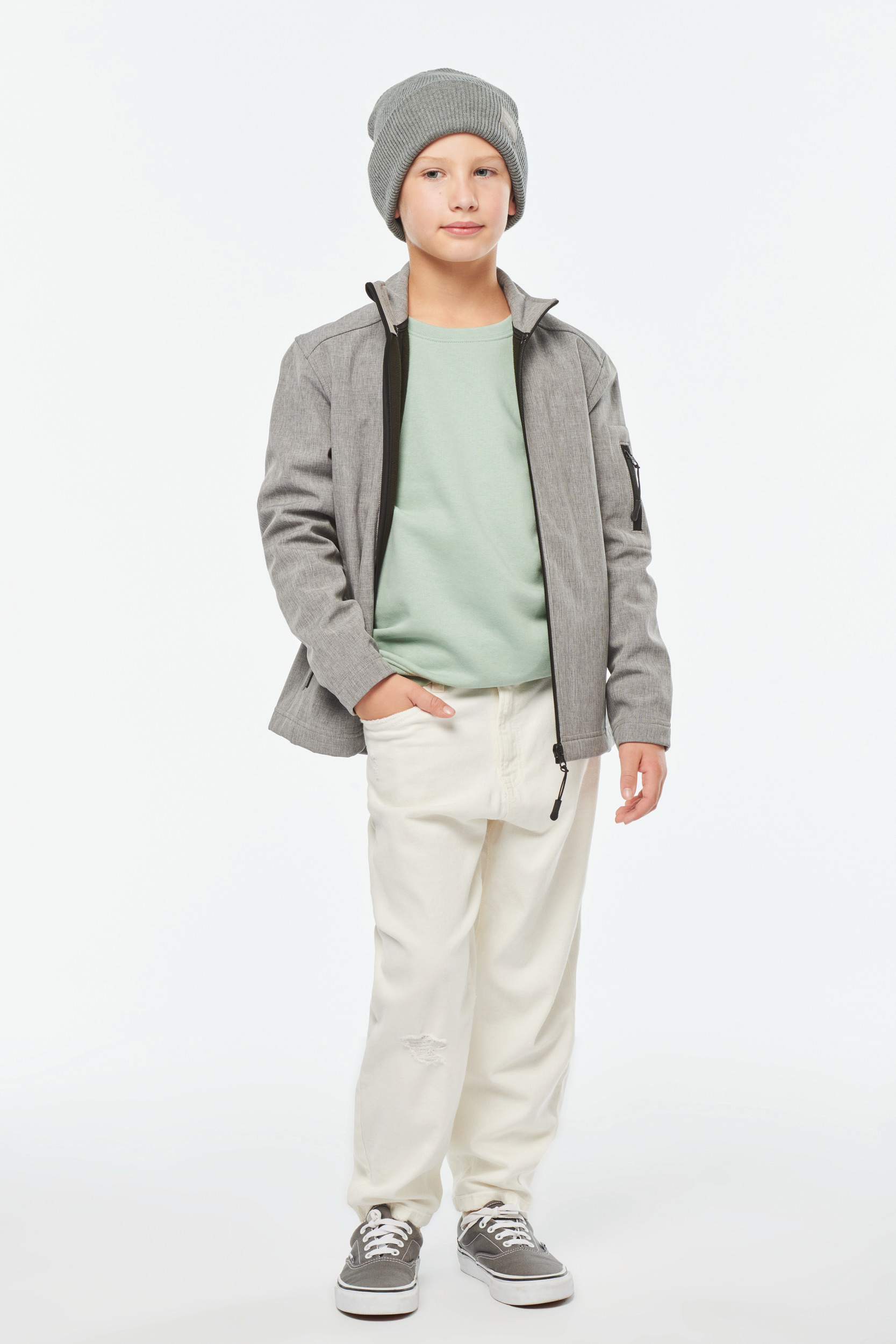 niño con gorro gris y chaqueta a juego, camiseta verde clara y vaqueros blancos de pie sobre fondo neutro
