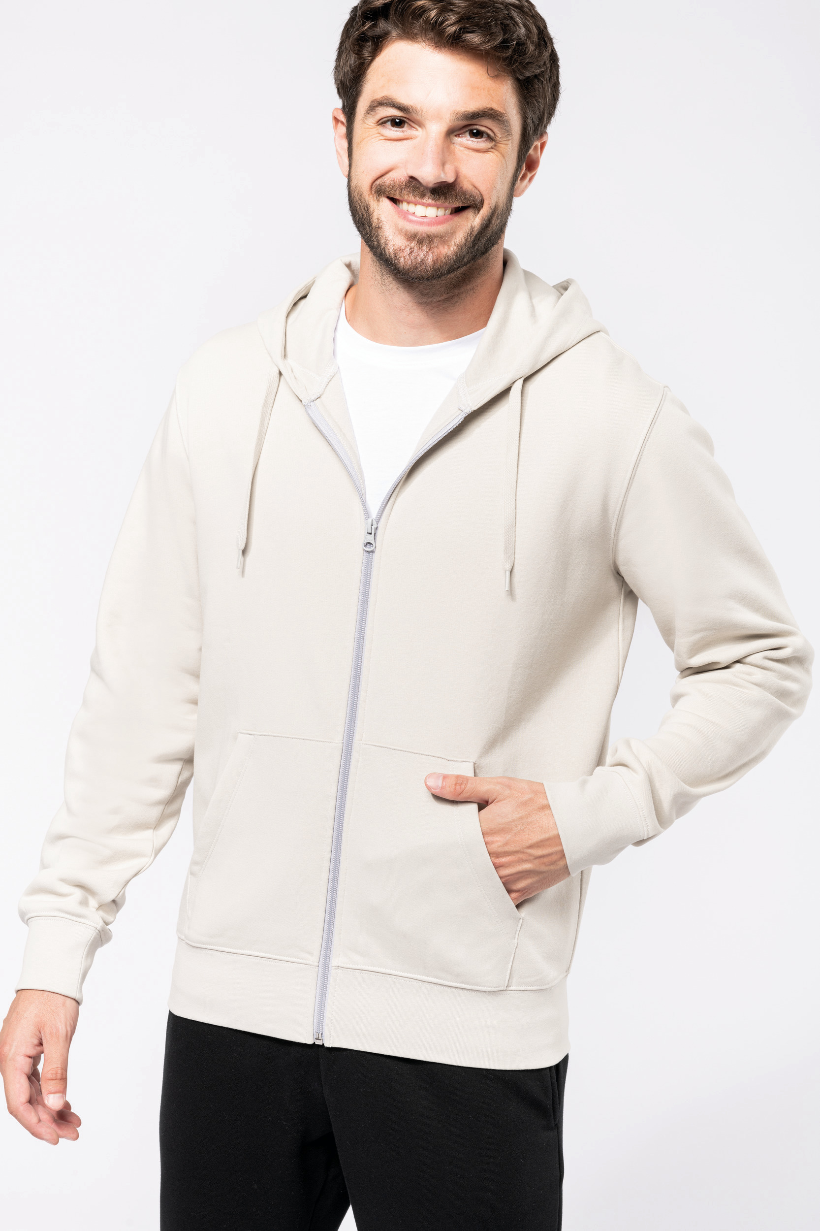 hombre joven sonriente con sudadera beige de cremallera y capucha con la mano en el bolsillo delantero