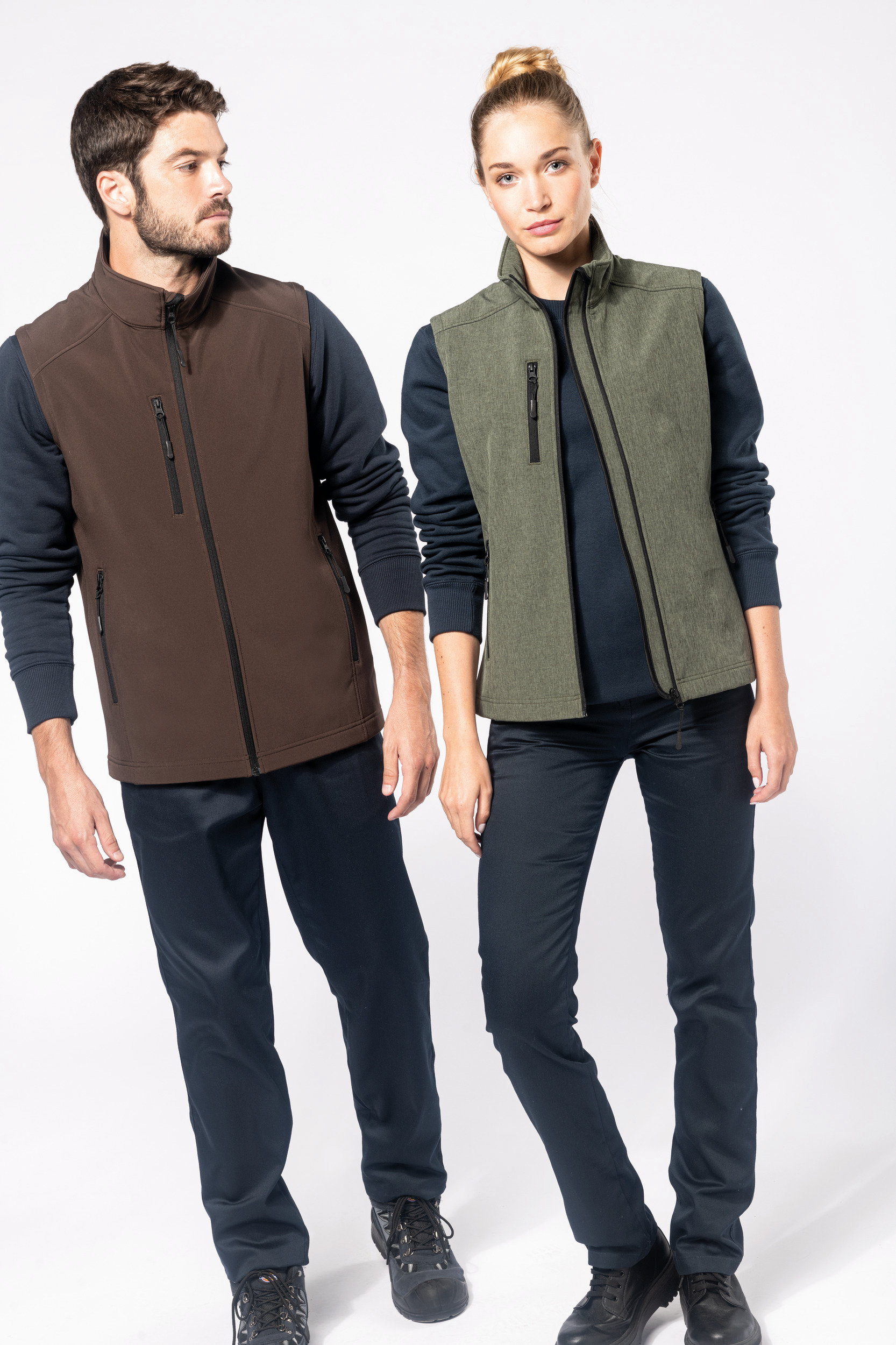 Hombre con chaleco marrón y mujer con chaleco verde posando con ropa informal sobre fondo blanco.