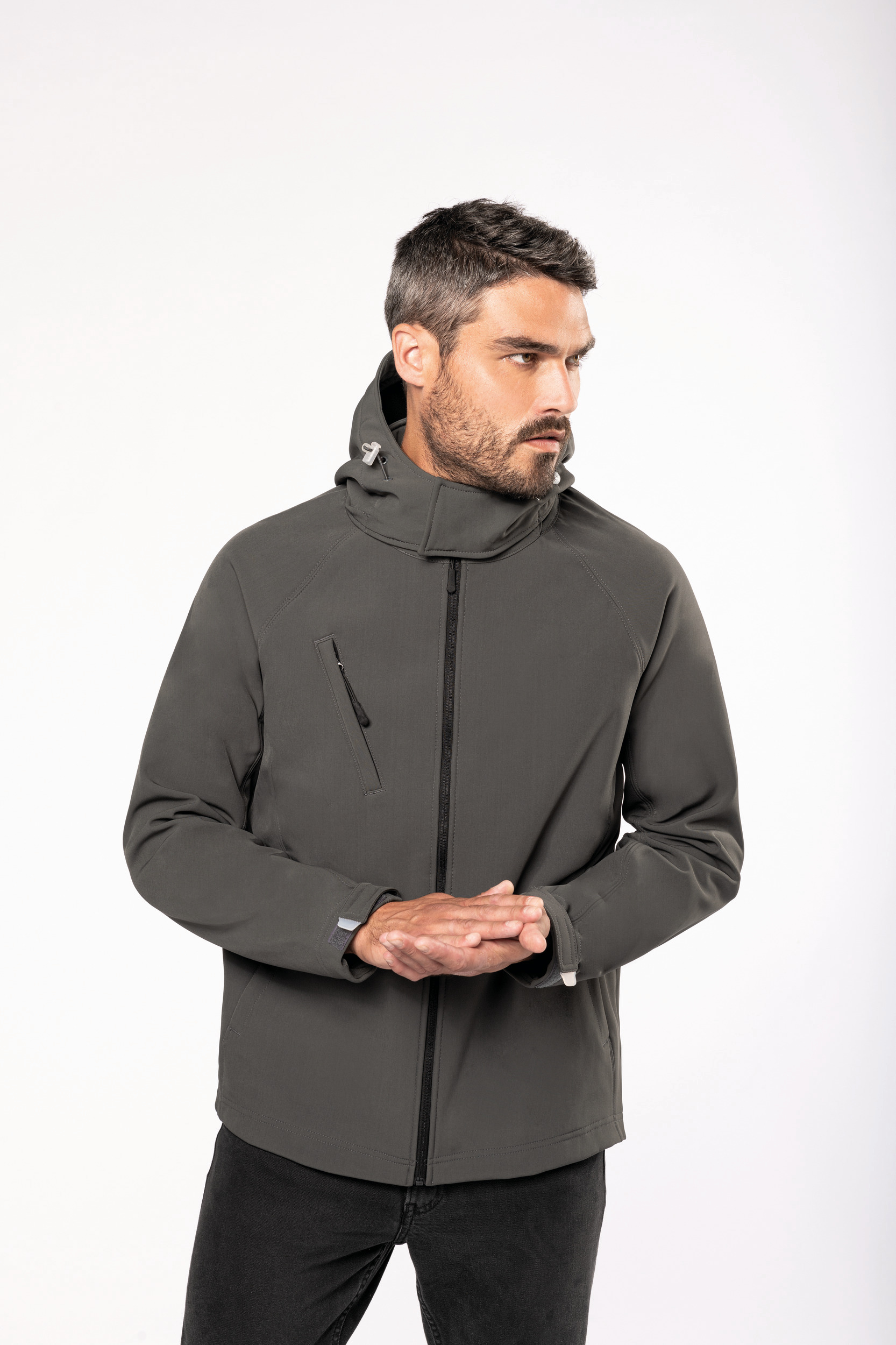 Hombre con chaqueta softshell gris oscuro con capucha y cremallera frontal posando de perfil con las manos juntas.