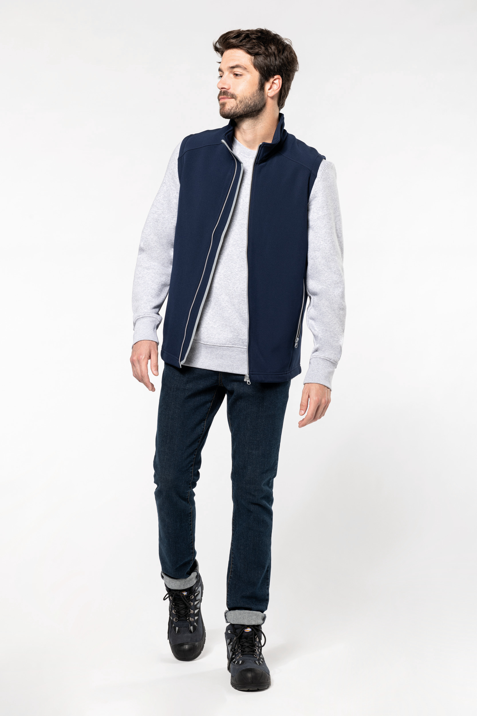 Hombre con chaleco polar azul marino sobre sudadera gris y vaqueros caminando sobre fondo blanco