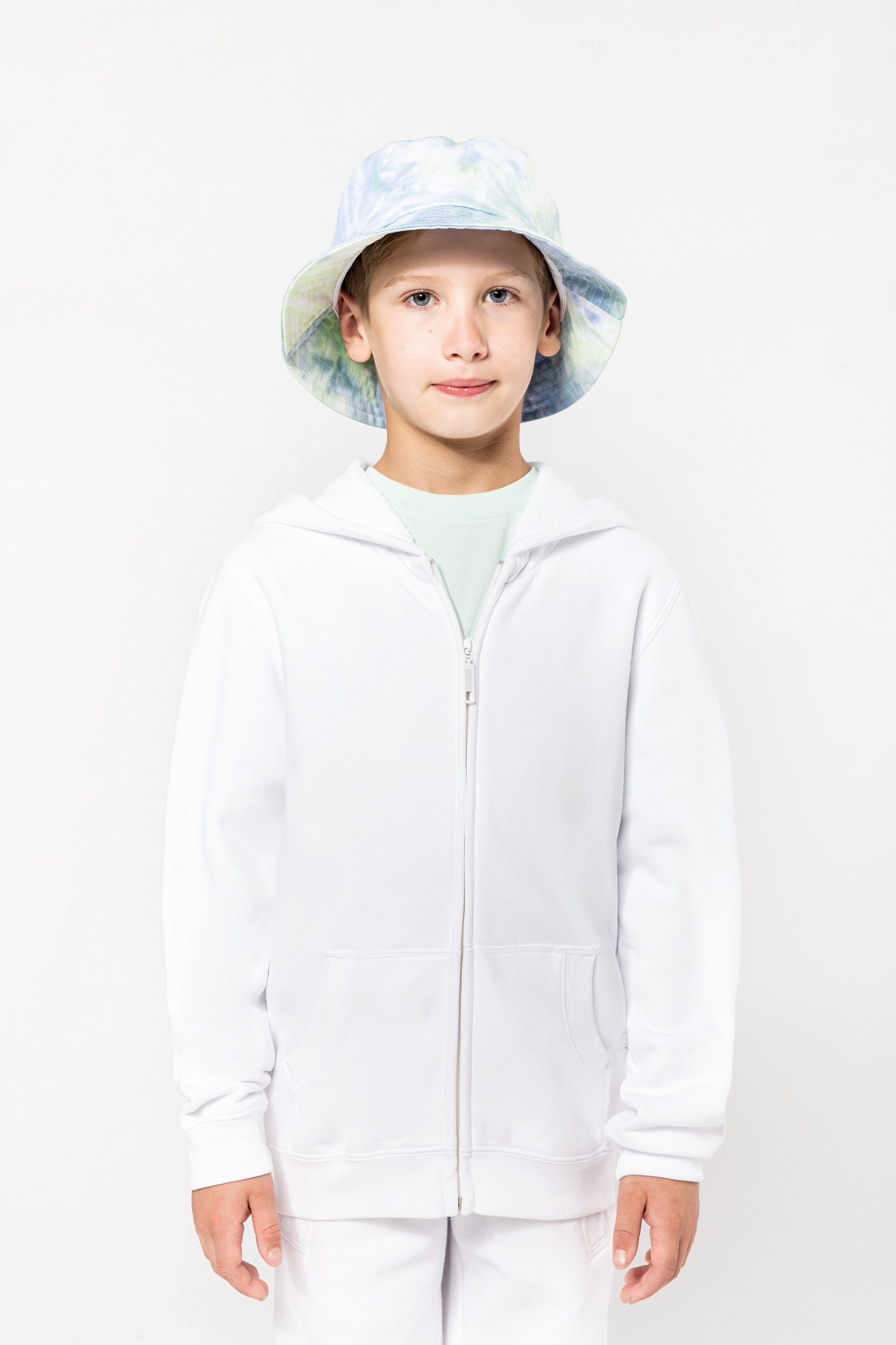 Niño de pie con sudadera blanca con cremallera y sombrero tipo bucket de estampado pastel sobre fondo blanco.