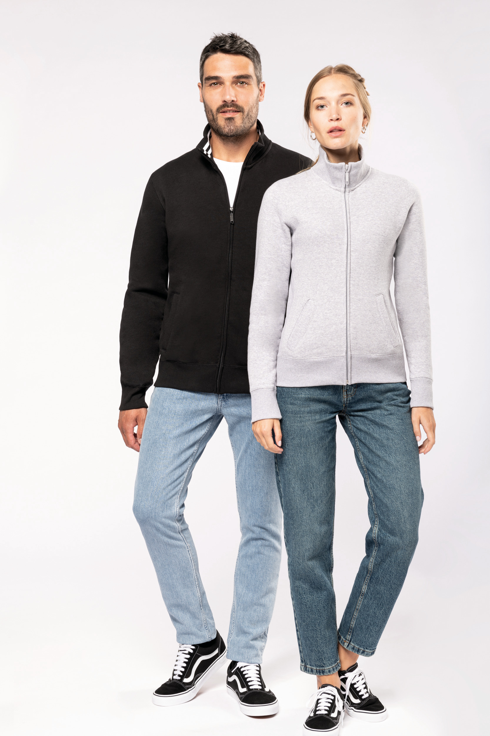 Hombre y mujer de pie con sudaderas de cremallera, vaqueros y zapatillas en fondo blanco.