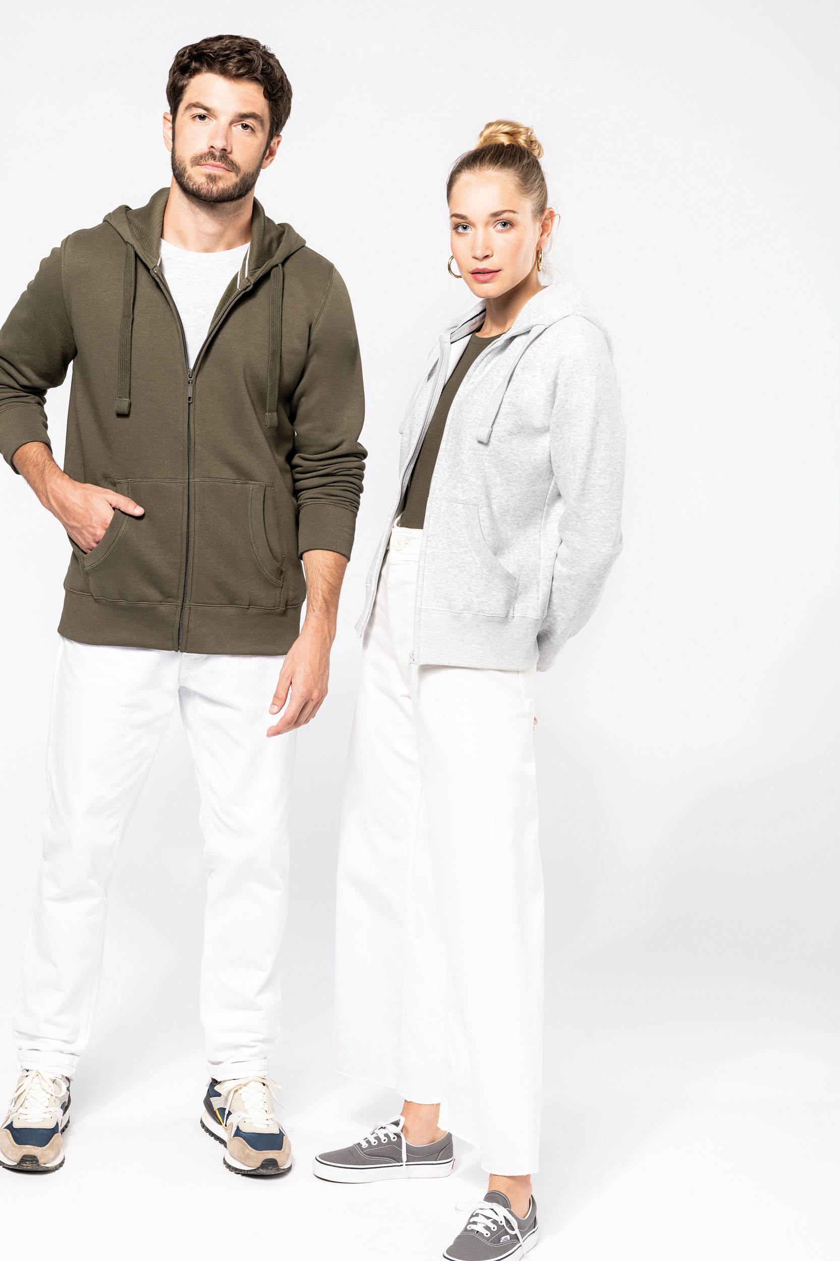 Hombre y mujer de pie con sudaderas con capucha y pantalones claros sobre fondo blanco.