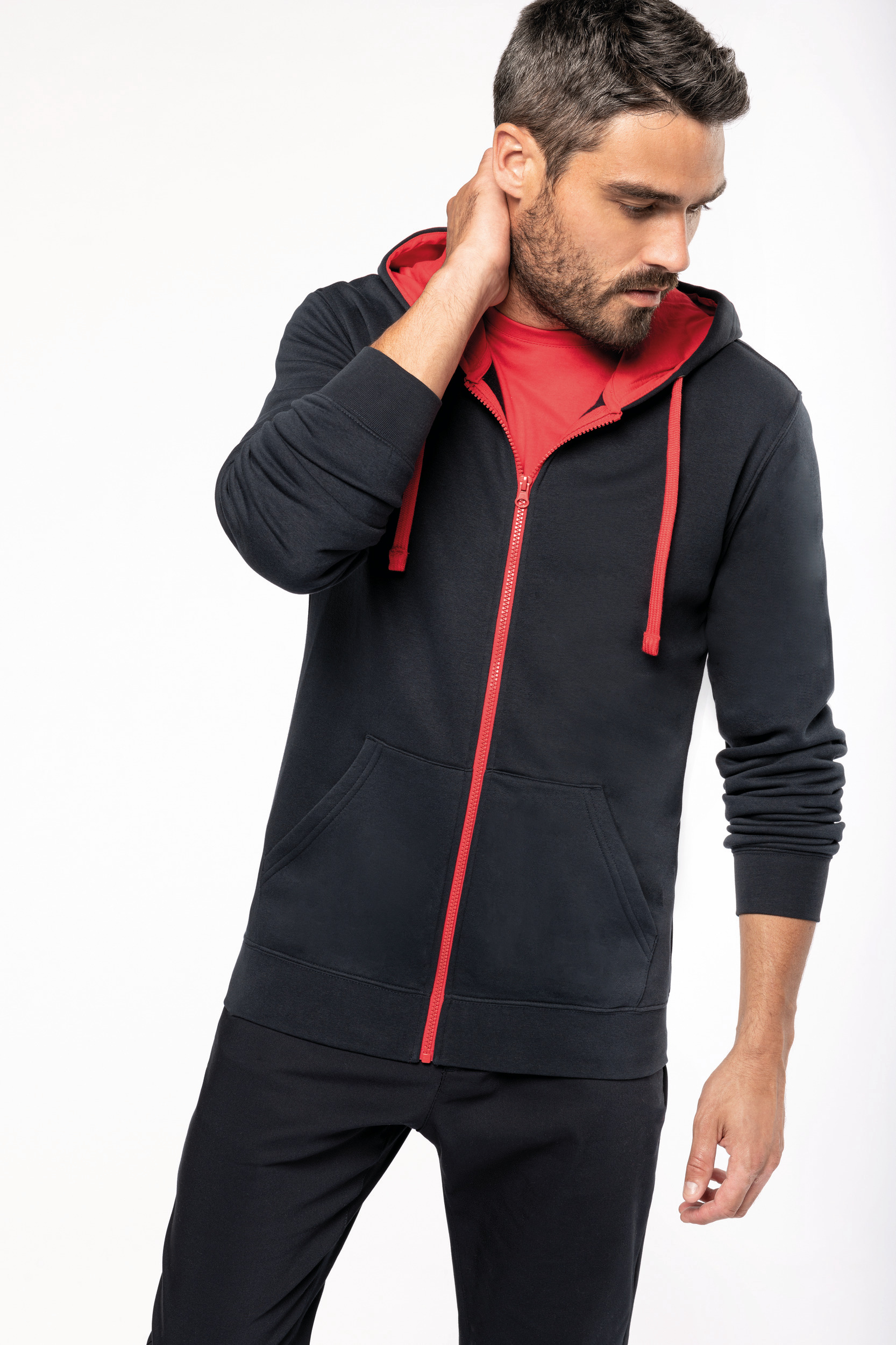 Hombre con sudadera negra de cremallera roja y capucha roja, posando con la mano en el cuello.
