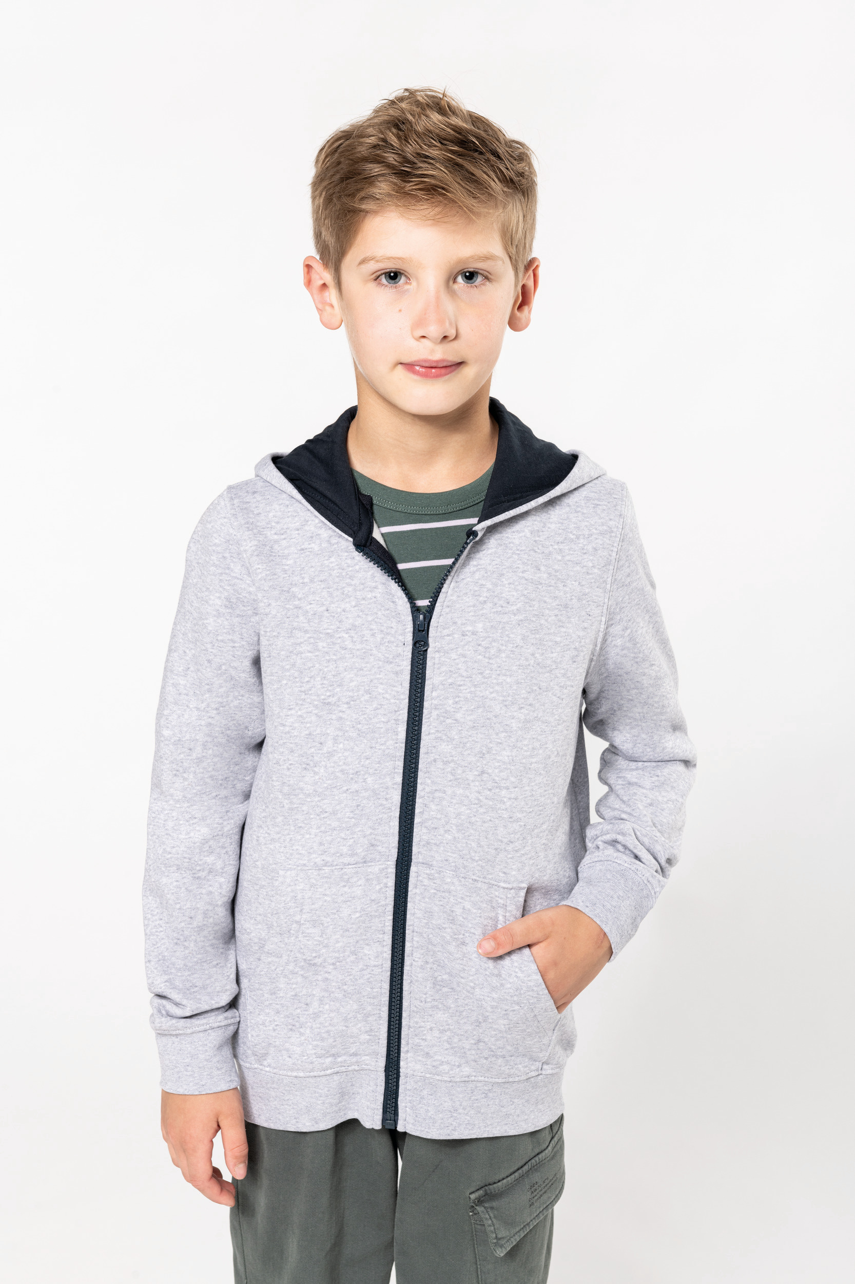 Niño de pie sobre fondo blanco con sudadera gris con cremallera y pantalones verdes, con una mano en el bolsillo.