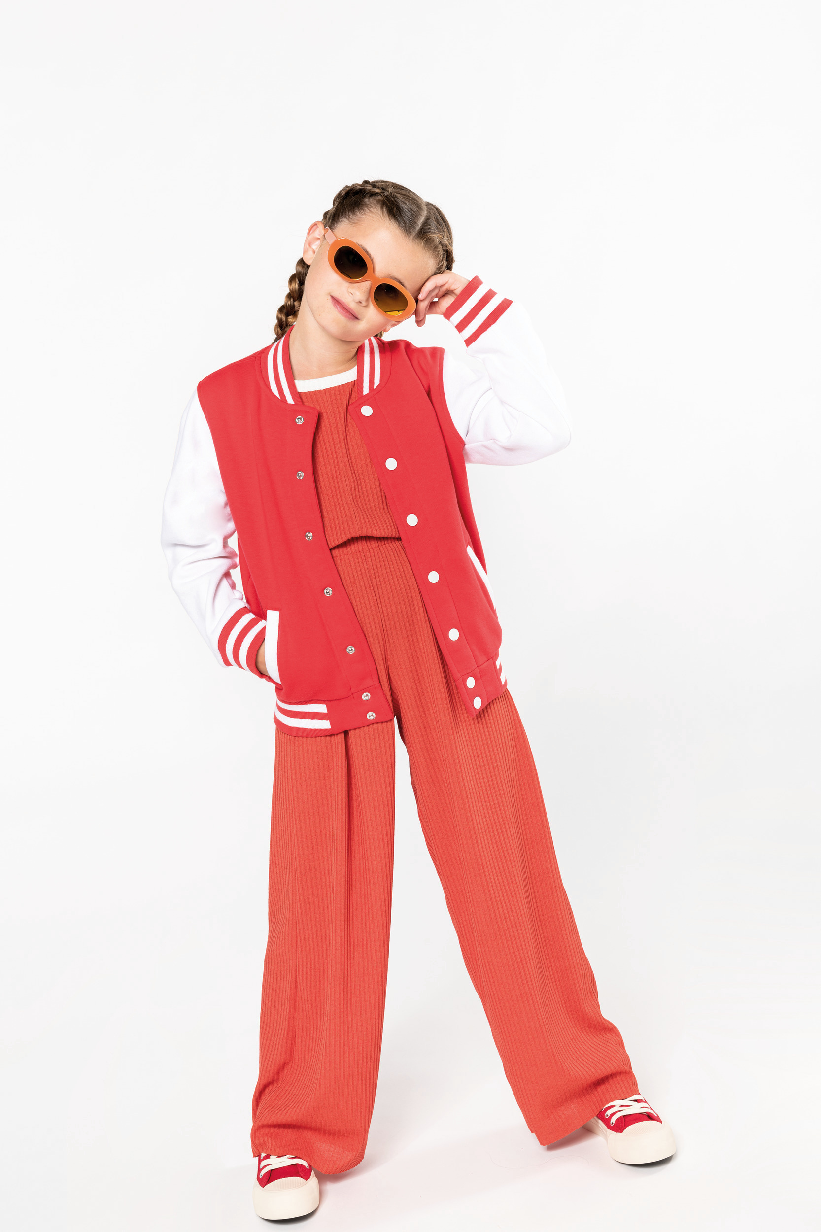 Niña de pie con chaqueta varsity roja, pantalones anchos naranja y gafas de sol naranjas sobre fondo blanco