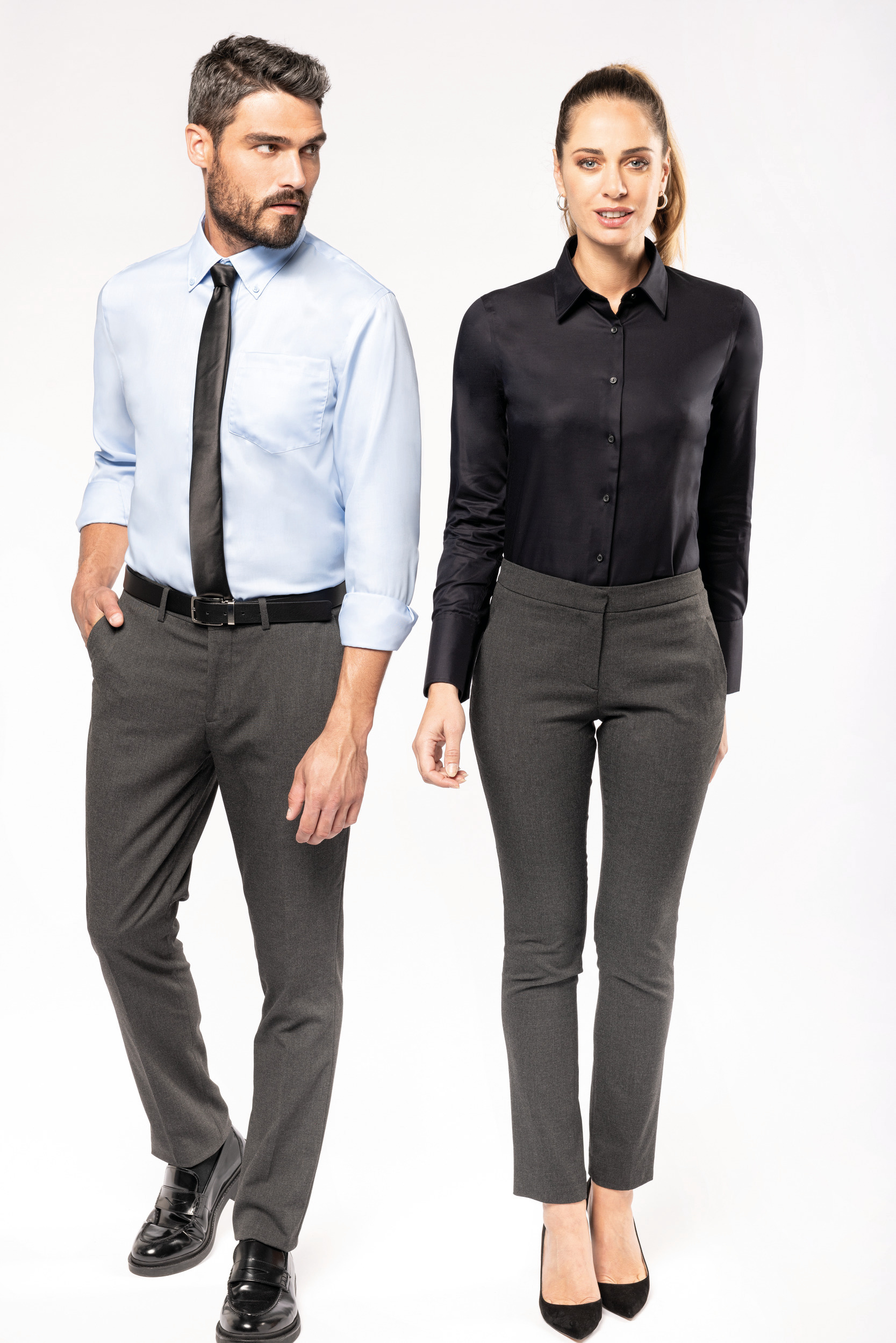 Hombre y mujer posando en fondo blanco con camisa de vestir y pantalón gris elegante.