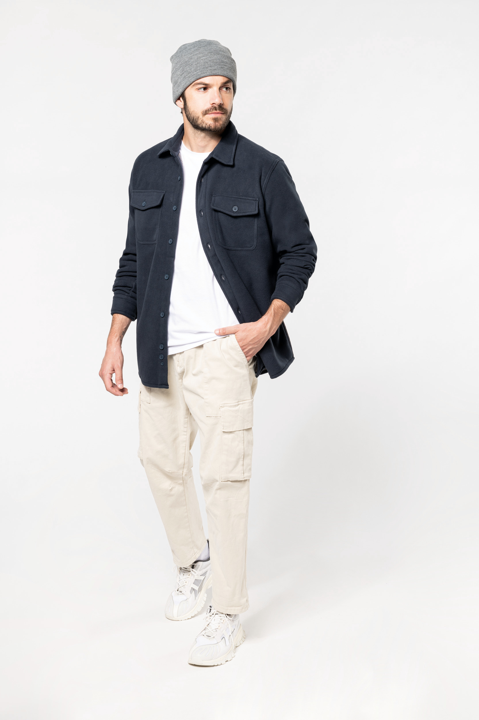Hombre con gorro gris, camisa overshirt azul marino, camiseta blanca, pantalones cargo beige y zapatillas blancas sobre fondo blanco