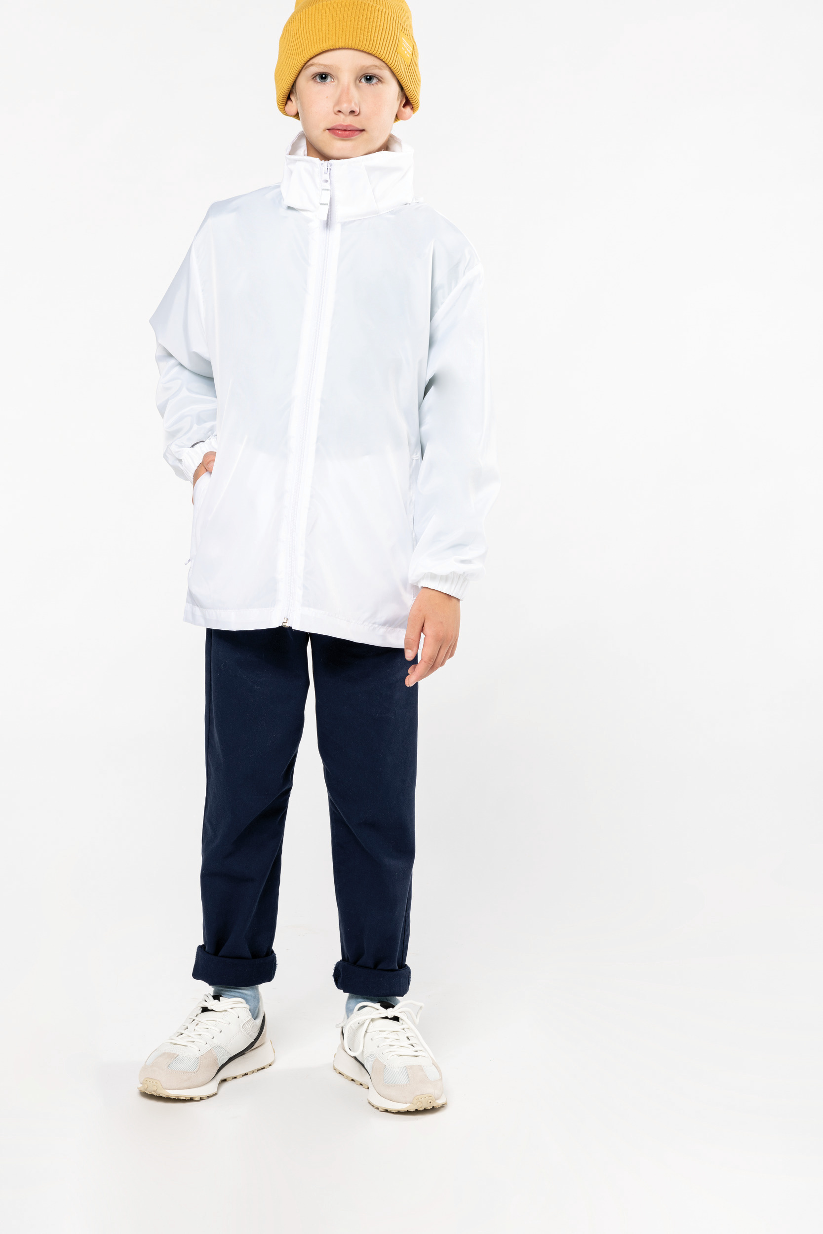 Niño de pie con chubasquero blanco, pantalones azul marino, zapatillas claras y gorro amarillo sobre fondo blanco