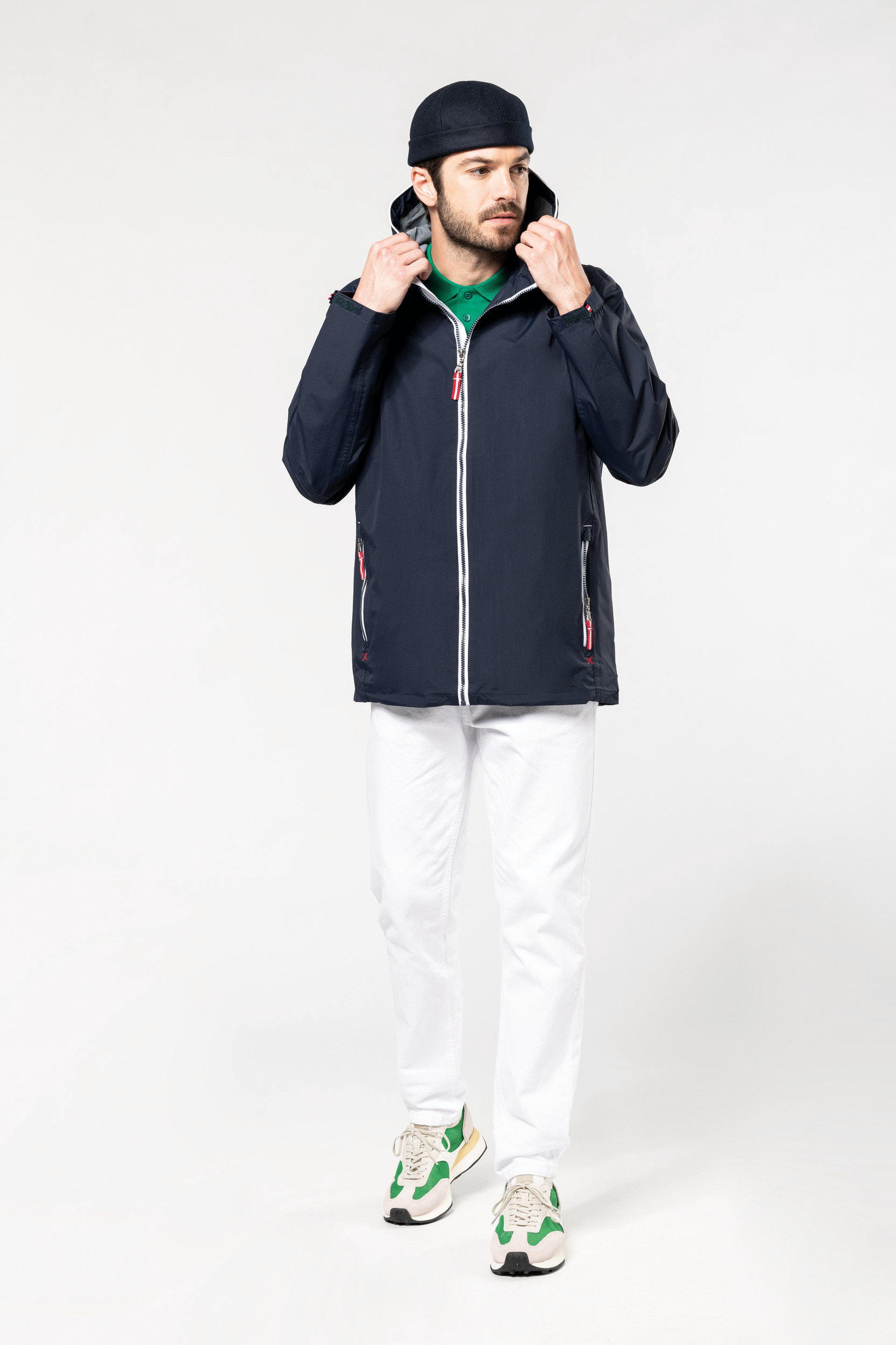 Hombre con chaqueta azul marino con capucha, polo verde, pantalones blancos y zapatillas verdes sobre fondo claro