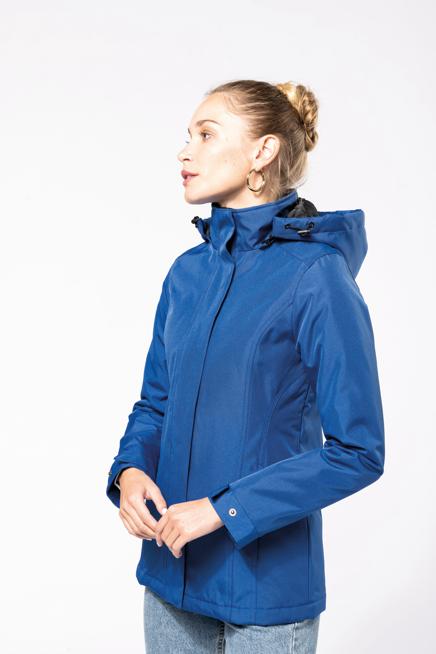 Mujer de perfil con chaqueta impermeable azul con capucha y vaqueros sobre fondo blanco.