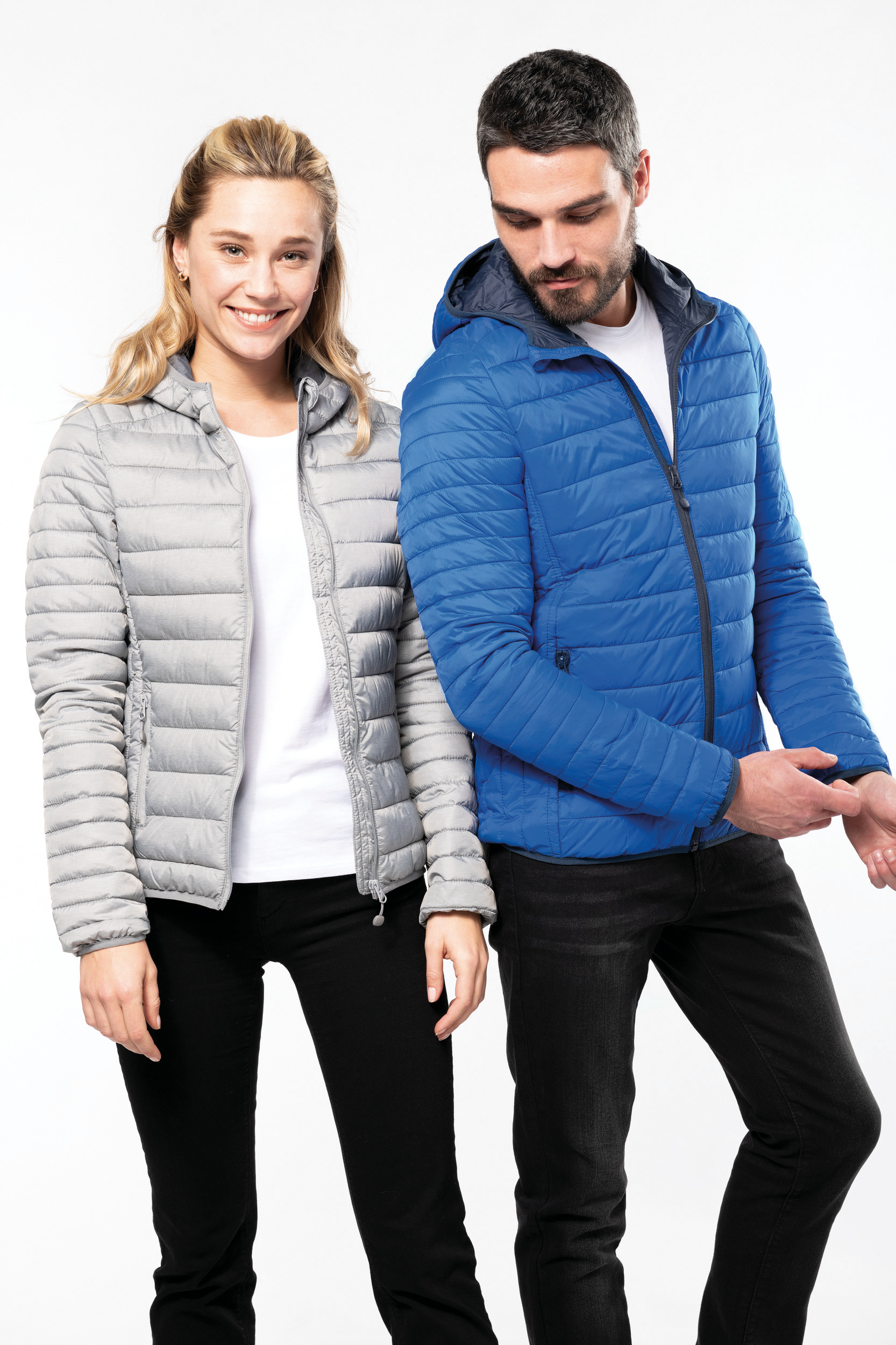 Mujer sonriente con chaqueta acolchada gris y hombre ajustando la manga de su chaqueta acolchada azul sobre fondo blanco
