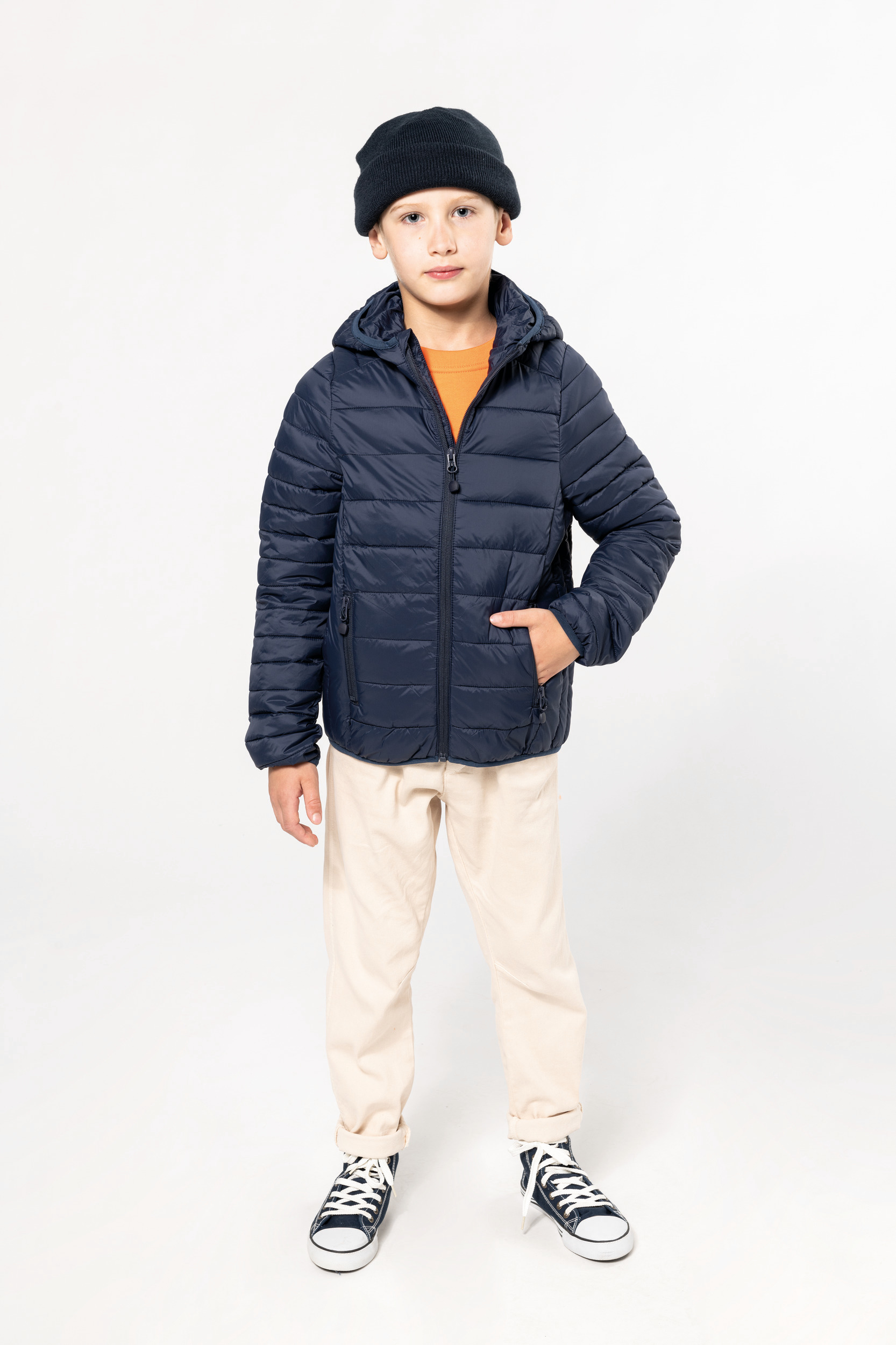 Niño de pie con chaqueta acolchada azul marino, gorro negro, camiseta naranja y pantalones claros.