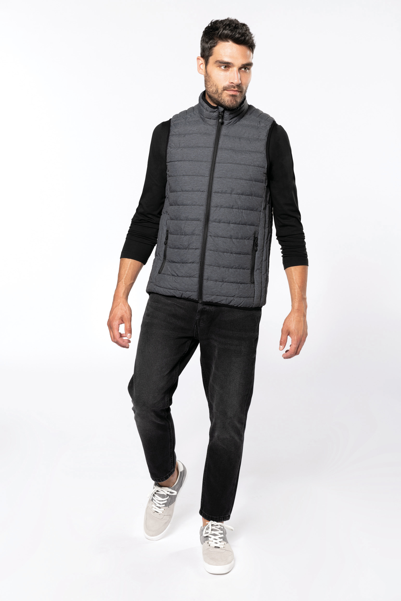 Hombre caminando en estudio con chaleco acolchado gris, camiseta negra y vaqueros oscuros